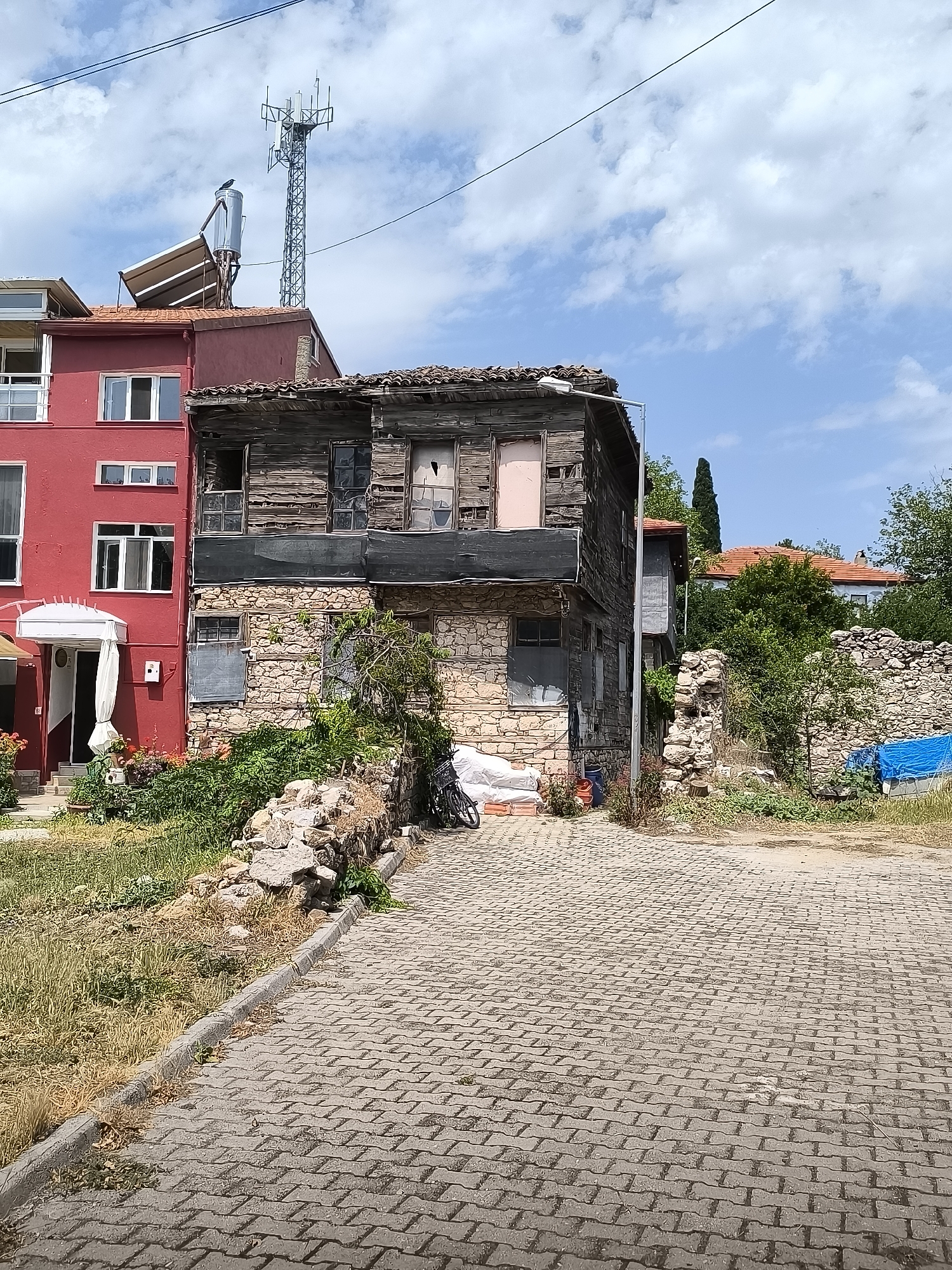 This image depicts a street scene in Eğirdir, Turkey, featuring a stark contrast between an old, dilapidated wooden and stone building and a modern, red multi-story structure. The old building, situated to the right, shows significant decay with weathered wooden planks, broken windows, and a crumbling stone foundation. In front of it, a patch of dry grass and scattered rocks are visible. Adjacent to this, a modern red building with clean white-framed windows and a white awning over an entrance stands prominently. A cell tower and a solar water heater are visible on the rooftop of the red building, indicating a more contemporary infrastructure. The foreground is dominated by a paved cobblestone walkway or street. The background reveals lush green trees and other buildings with terracotta roofs, suggesting a residential or mixed-use area. The sky is partly cloudy with blue patches. No people are present in the frame, and no actions or interactions are depicted. The scene appears to be an outdoor, daytime setting.