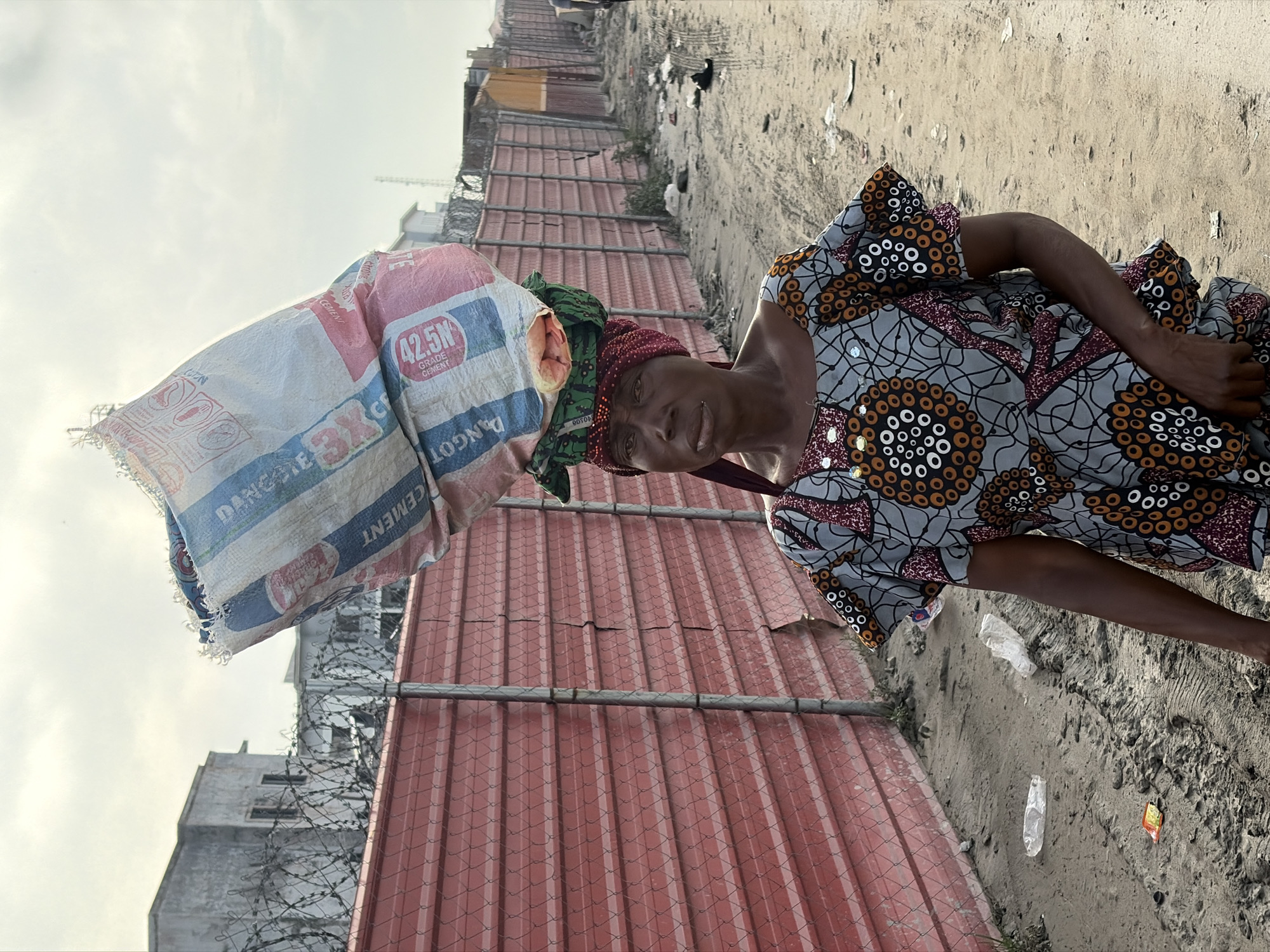 A medium shot captures an adult African woman with dark skin and short, dark hair, wearing a colorful, patterned dress and a red headscarf, carrying a large, bulging bag of cement on her head. The bag, a large white and blue sack, appears to be for cement, with visible red and blue lettering. She is standing on a dirt path in what appears to be a construction or developing area. Behind her, a vibrant red corrugated metal fence stretches across the frame, topped with barbed wire. Beyond the fence, partially visible buildings with concrete facades and windows can be seen, along with a cloudy sky. The ground is sandy and appears somewhat uneven, with tire tracks and scattered debris, including plastic bottles and wrappers. The overall scene suggests an outdoor, possibly industrial or semi-urban environment in Alaguntan, Nigeria. The woman's expression is serious as she looks directly at the camera, her body turned slightly to the left.