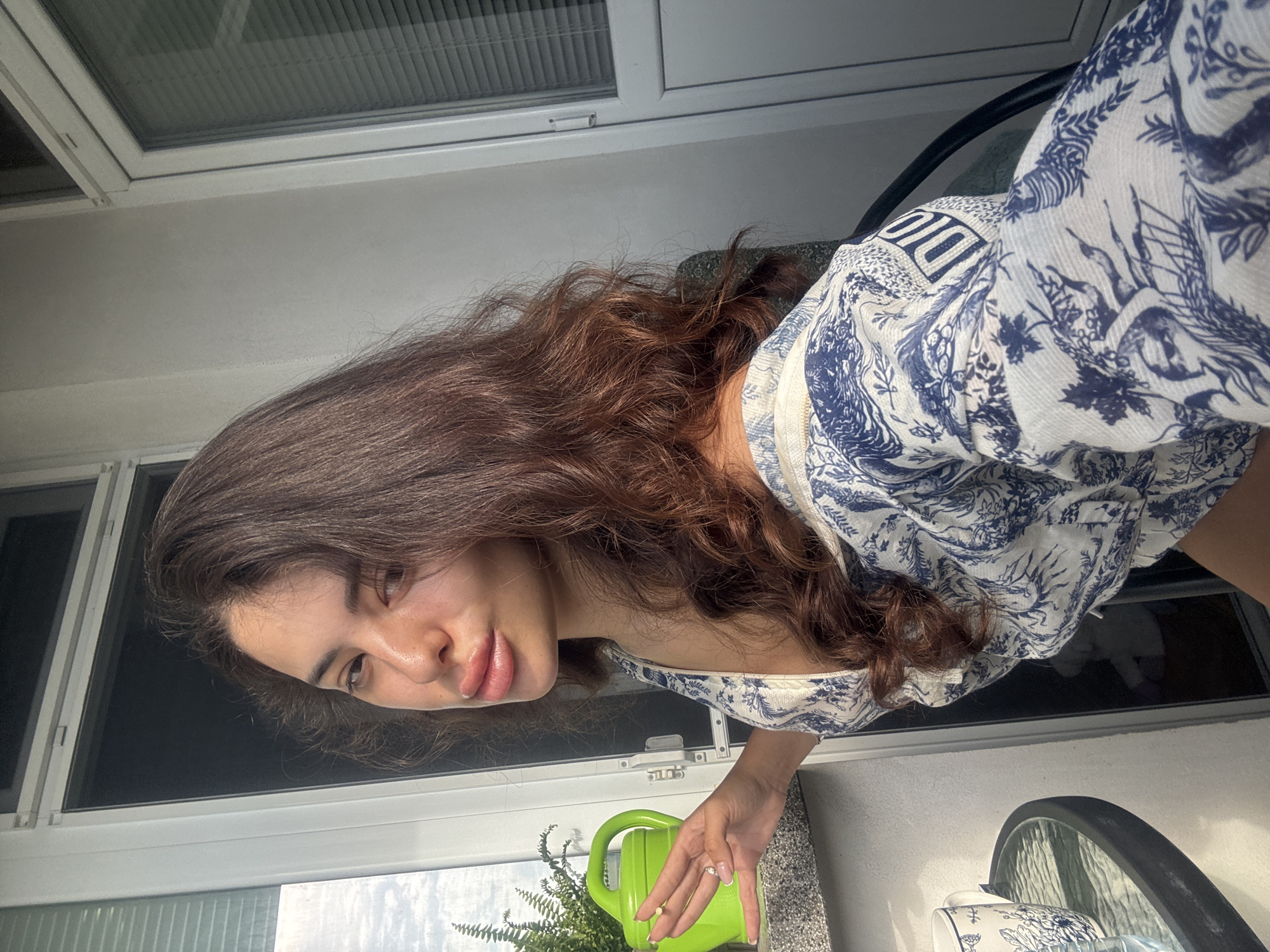 A young woman with long, dark, wavy hair and full lips is leaning against a balcony railing, holding a small, green watering can. She is wearing a white and blue patterned dress. A potted fern is visible behind her. The balcony appears to be part of an apartment building in Plovdiv, Bulgaria. The lighting suggests it is daytime.