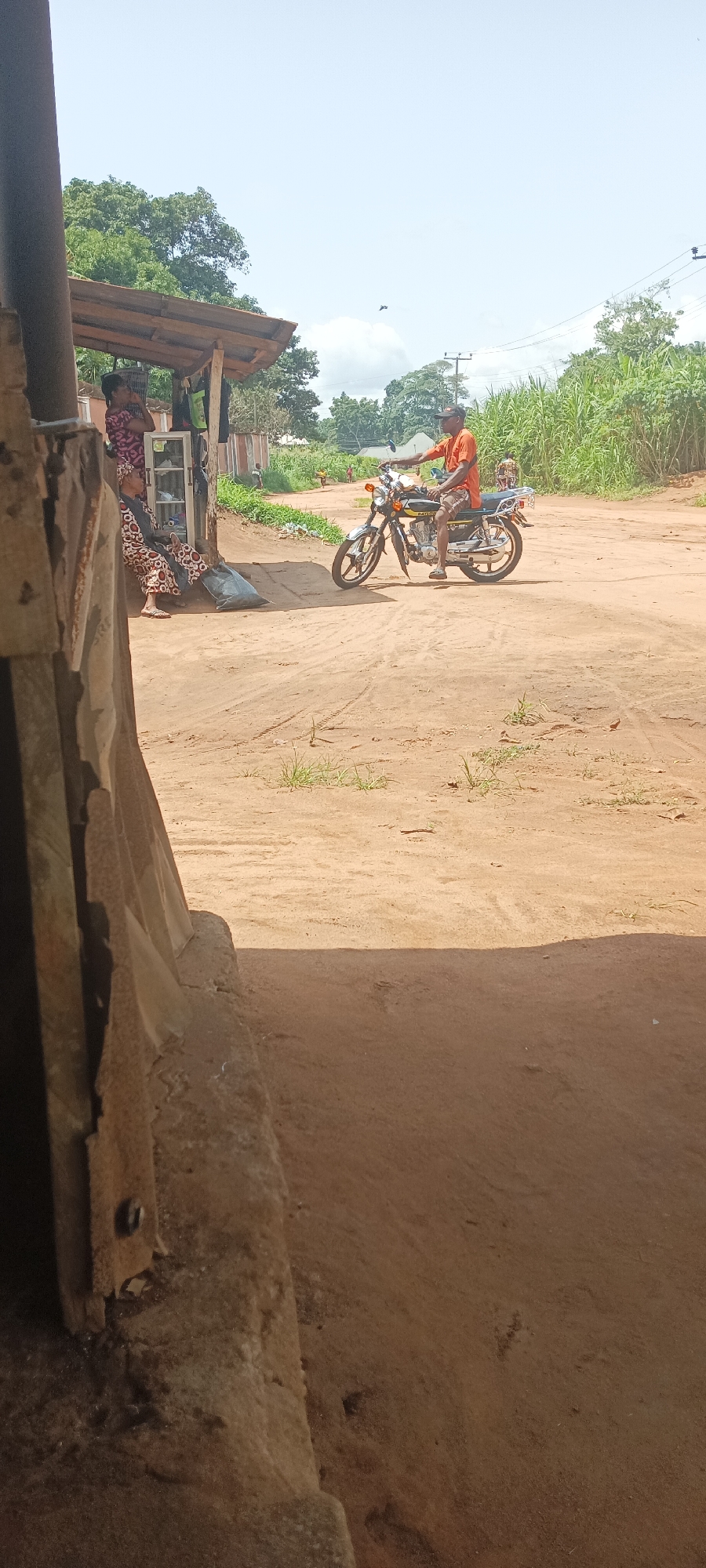 The scene captures a dusty street corner in what appears to be a rural area near Amauzu, Nigeria. A man in an orange shirt is sitting on a motorcycle, positioned in the middle of the sandy road. Near him to the left, under a makeshift wooden shelter, two women are engaged in what seems to be a conversation. In the background are scattered houses, vegetation, and power lines. The foreground is dominated by a weathered, concrete structure, obscuring a portion of the scene, suggesting the image was taken from inside a building.