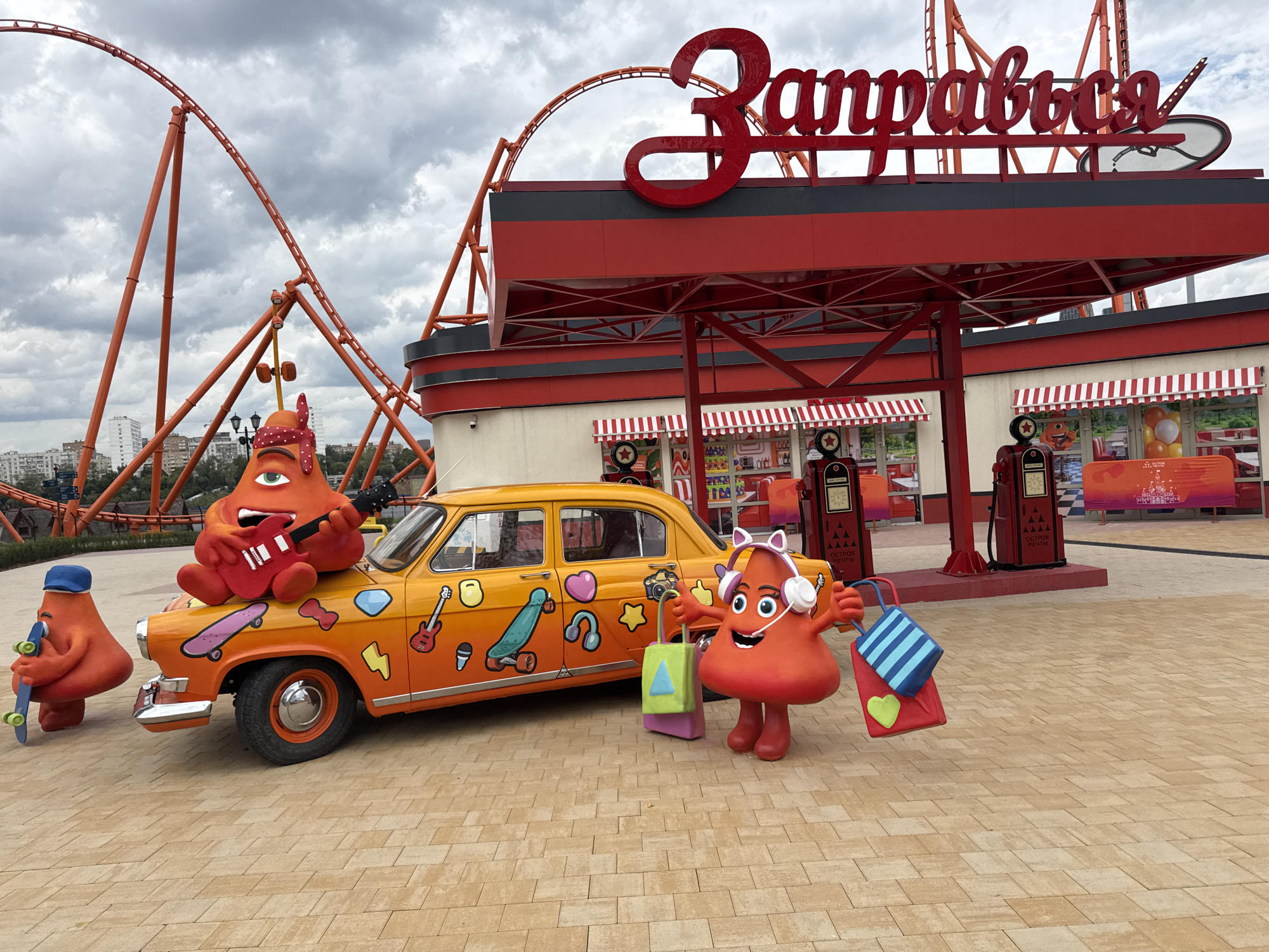 In Moscow, Russia, at the "Zapravisa" amusement park, an orange retro car is parked on a beige paved area. The car is decorated with various colorful stickers, including guitars, headphones, and skateboards. A large, red, cartoonish character resembling a stylized tomato with a single eye and a red bandana sits on the car's hood, playing a red electric guitar. To the left of the car, another red, cartoonish character, this one wearing a blue cap, holds a skateboard. To the right of the car, a third red, cartoonish character, this one wearing headphones and a bow, holds two shopping bags. In the background, a red roller coaster structure looms against a cloudy sky, and a red, retro-style service station with two gas pumps stands behind the car.