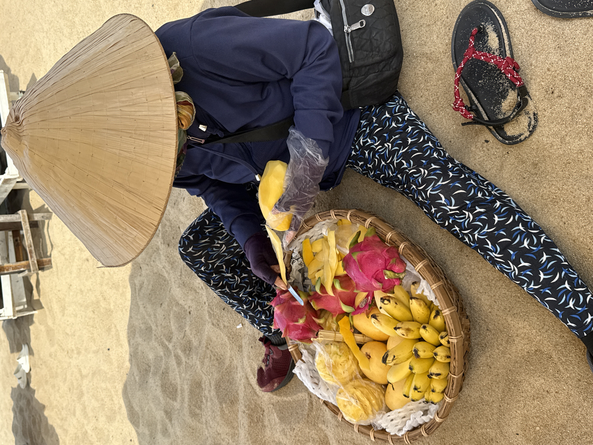 A high-angle, top-down shot captures a person sitting on a sandy beach, engaged in preparing tropical fruit. The individual is wearing a traditional Vietnamese conical hat (nón lá) that largely obscures their face. They are dressed in a dark blue long-sleeved top and dark patterned leggings with white and blue stylized motifs. A black backpack with a metallic emblem, seemingly bearing the brand name "REVERIE," rests beside them on the sand.
The person is holding a piece of yellow fruit, likely a mango or similar, in a hand covered by a clear plastic glove, and using a knife in the other hand to peel or cut it. A large, round woven basket sits in front of them, filled with a vibrant assortment of tropical fruits, including bright pink and green dragon fruits, numerous ripe yellow mangoes, and small clusters of bananas. Some fruit peels and prepared fruit pieces are also visible within the basket.
The setting is a sunny beach, with soft golden sand and clear shadows indicating daytime. A pair of black flip-flops with red knotted straps are visible on the sand near the person's leg. In the background to the left, light-colored wooden structures are partially visible. The scene depicts a quiet moment of fruit preparation, characteristic of beachside vendors, in Nha Trang Ward, Vietnam.