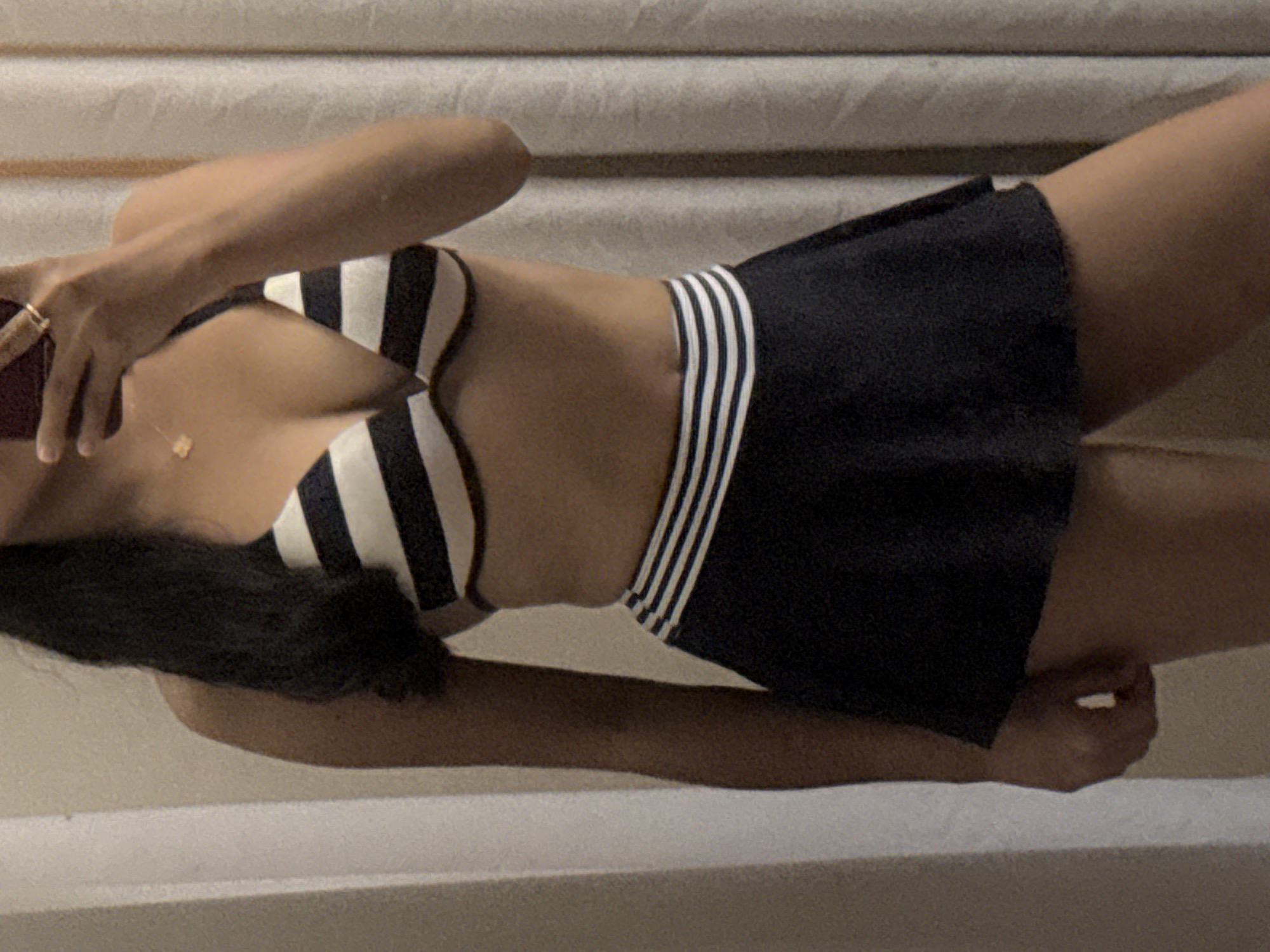 The image displays a waist-up and upper-thigh view of an individual taking a mirror selfie indoors. The person, appearing to be female, has medium brown skin and long, dark hair styled over her left shoulder. She is wearing a two-piece swimwear set consisting of a black and white horizontally striped halter-neck bikini top and a black swim skirt with three thin white horizontal stripes at the waistband. Her right hand, adorned with a gold ring on the ring finger, holds a dark-colored smartphone, which obscures her face. A delicate gold necklace with a small pendant is visible around her neck. The background on the left features a light-colored, possibly off-white, textured wall or vertical architectural element. On the right, a light-colored, vertically pleated curtain is visible. The specific location of Davao City, Philippines, cannot be determined from the visual information provided in the image.