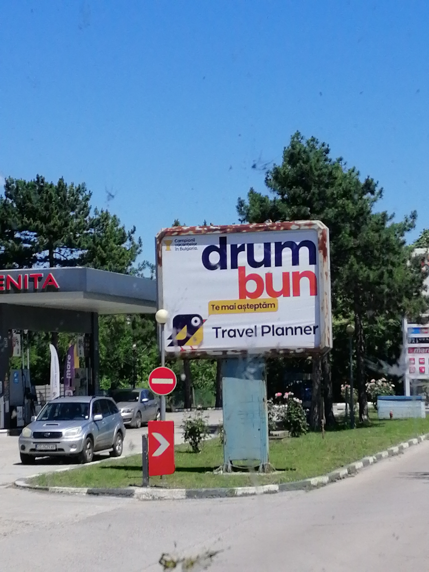 The image depicts a sunny day scene at a gas station. In the foreground, a silver SUV is parked to the left of the frame. Next to it, a red directional sign with a white arrow points to the right. Further in the background, a large billboard stands prominently. The billboard features text that reads "drum bun" in large, bold letters, with "bun" in red. Below this, "Te mai aşteptăm" is written, followed by "Travel Planner". A small, cartoonish bird with yellow and black coloring is also visible on the billboard. To the left of the billboard, a gas station canopy with the red lettering "ENITA" is partially visible. The surrounding environment consists of green grass, trees, and a clear blue sky. There are no people clearly visible in the image.