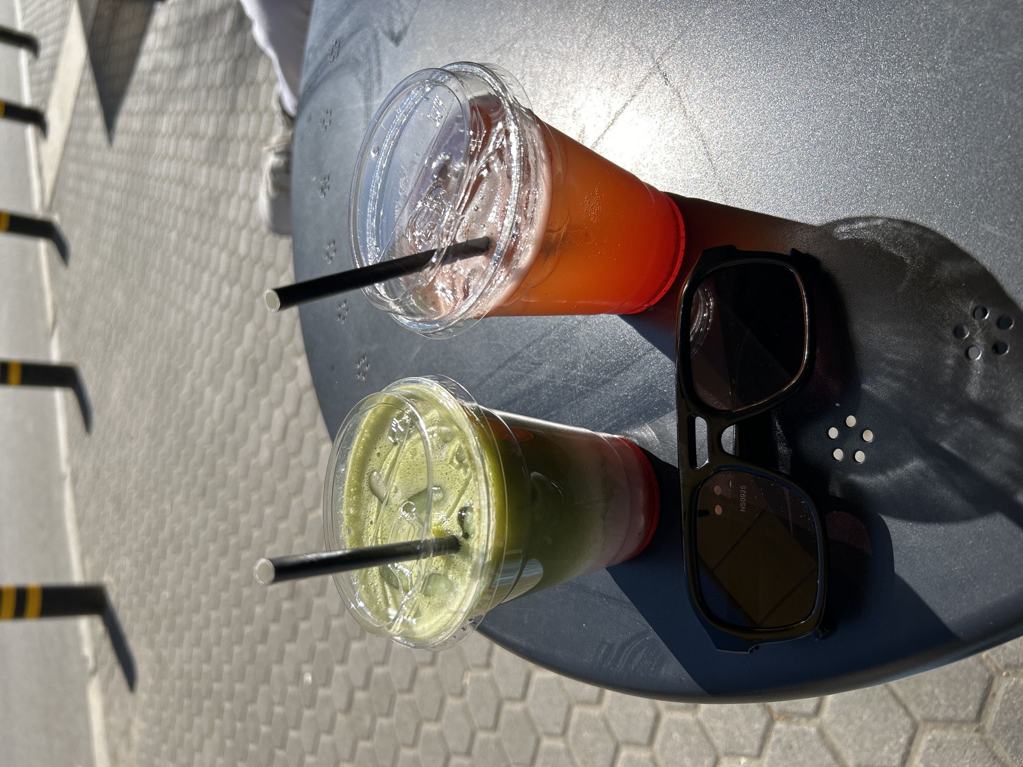 The image shows two plastic cups with beverages and a pair of sunglasses on a dark gray, possibly metallic table, likely outside. One cup contains a reddish-orange liquid with ice and a black straw. The other cup has a layered drink with green on top and red below, also with ice and a black straw. The sunglasses are black-framed. The table appears to be in a public area, possibly a cafe or outdoor seating area, as indicated by the paving stones and a partially visible railing with yellow and black markings. The setting is likely Sofia, Bulgaria, as the image was captured in Sofia, Bulgaria.