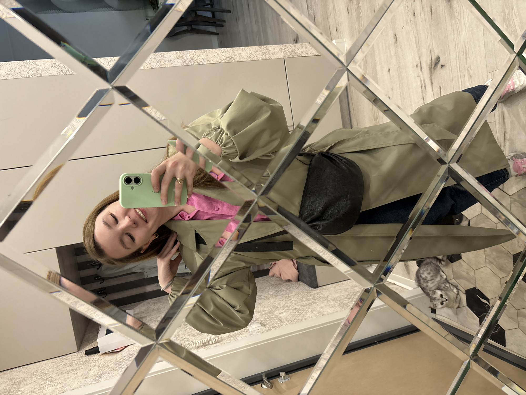 A woman is visible in the reflection of a wall mirror composed of diamond-shaped tiles. She is holding a green smartphone, presumably taking a selfie. She is wearing a pink shirt under an olive green trench coat. A black crossbody bag is slung across her body.
The floor visible in the reflection is covered with a light-colored rug or carpet. A small grey tabby cat is standing on a floor with black and white hexagonal tiles. The reflection also shows glimpses of what appear to be a dresser, clothing, and interior architectural details.
The location appears to be indoors, possibly in a residence. There is no information available from the image itself to confirm that the location is in Izhevsk, Russia.