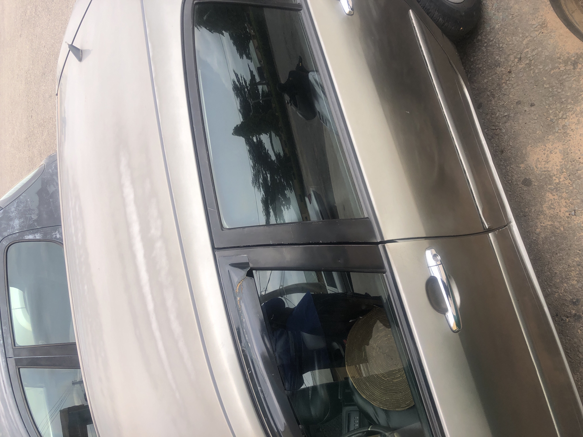 The image shows the side of a beige car parked on a gravelly surface in Jalingo, Nigeria. The car has tinted windows and a black trim around the windows. A part of another dark-colored car is visible in the upper left corner of the image. The beige car has a chrome door handle. Inside the car, a woven hat is visible on the passenger seat, and a dark blue fabric is draped over something. The reflection in the tinted windows shows trees and a body of water. A shark fin antenna is visible on the roof of the beige car. The overall environment appears to be an outdoor urban or semi-urban setting.