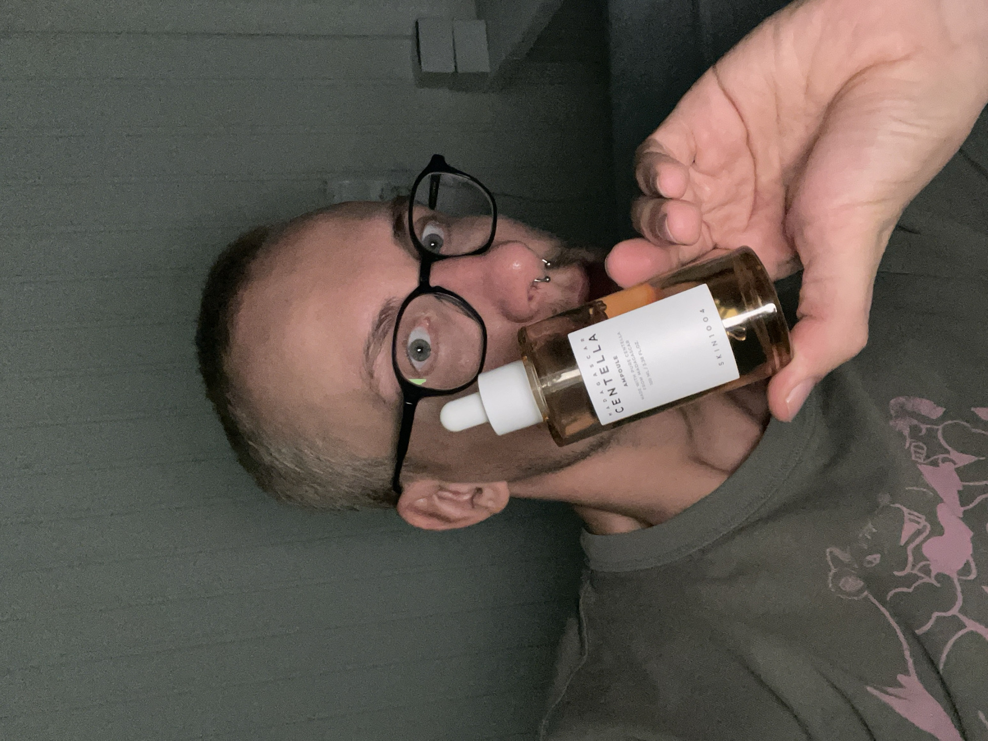 A man with glasses, a nose piercing, and short hair is holding a bottle of Madagascar Centella Ampoule by SKIN1004, appearing to place the dropper in his mouth. The man is wearing a t-shirt with a purple cartoonish design. The background appears to be a green textured wall. There is no evidence this image was taken in the city of Sestroretsk, Russia.