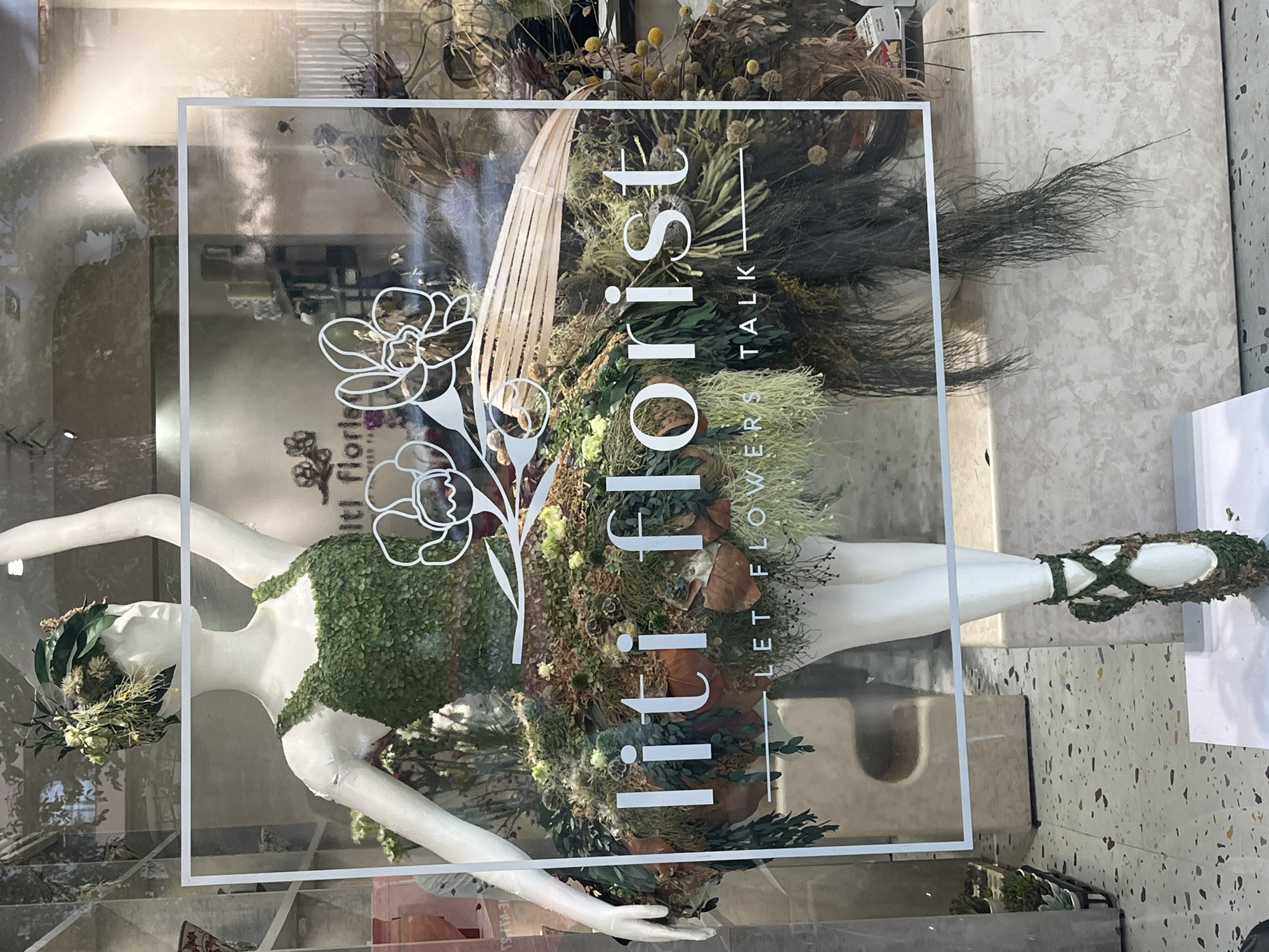 A mannequin dressed in a floral outfit, resembling a ballerina's costume, is displayed in a shop window. The mannequin has a white body and is adorned with various green leaves and dried flowers. The dress consists of a green leaf-covered bodice and a voluminous tutu made of moss and dried foliage. The mannequin's headpiece is also made of greenery. The shop window has text overlayed on it that reads "liti florist" and "LET FLOWERS TALK". The reflection of trees and the sky are visible on the glass. The background of the shop appears to be a florist's interior with shelves and floral arrangements. The floor in front of the display has a terrazzo pattern. The overall scene suggests a flower shop in Thủ Đức, Vietnam.
