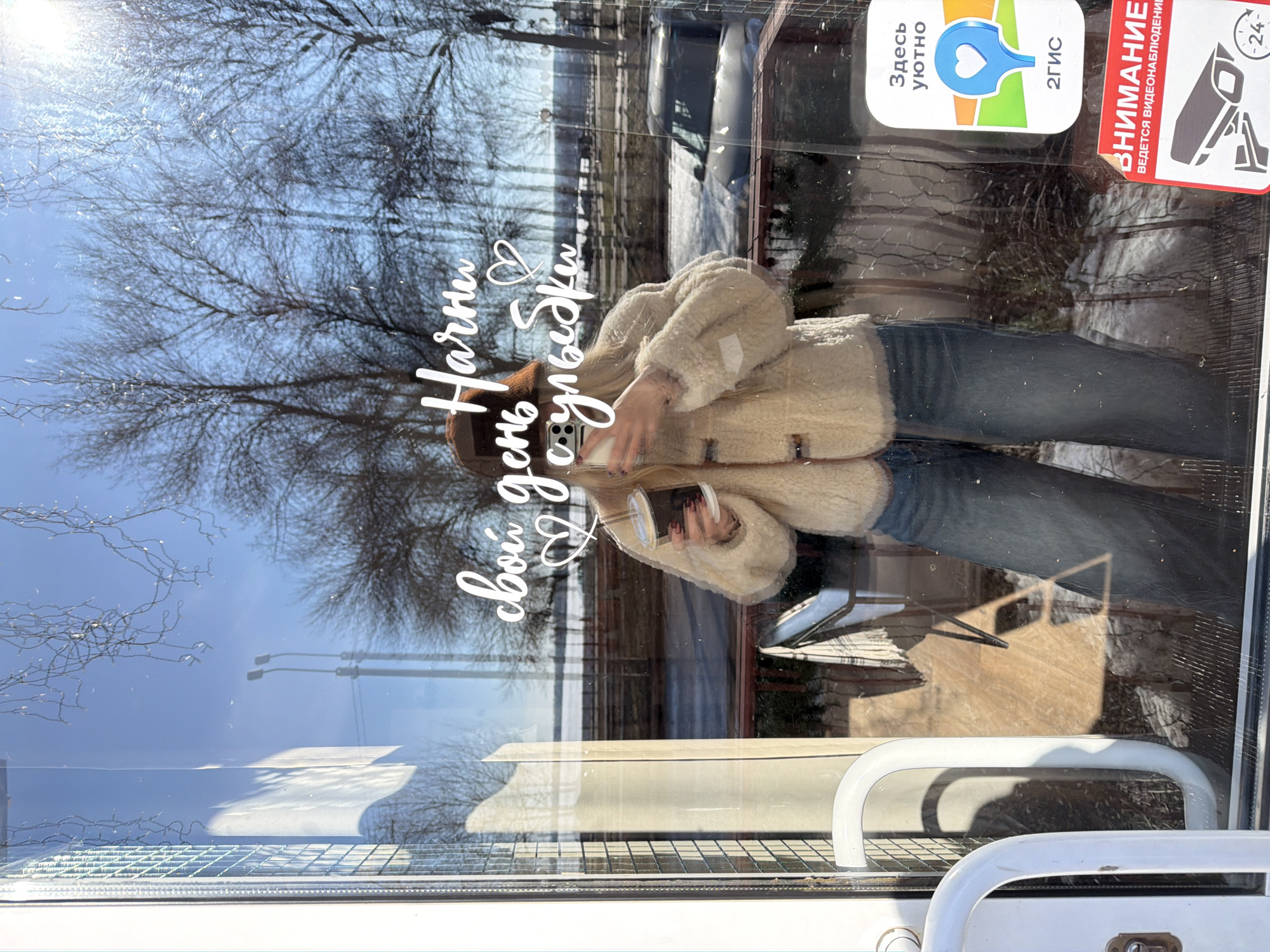 A woman is reflected in the window of a building. She is wearing a brown hat, a cream-colored jacket, and blue jeans. She is holding a smartphone. The window has Russian text in white lettering that reads, "Начни свой день с улыбки". A sticker with a blue and green logo and the word "Здесь уютно" is visible in the upper right corner. Another red sticker with a surveillance camera graphic is also in the upper right corner. The reflection shows trees without leaves and a building across the street. The handle of a door is visible at the bottom of the image. The building may be in Votkinsk, Russia.