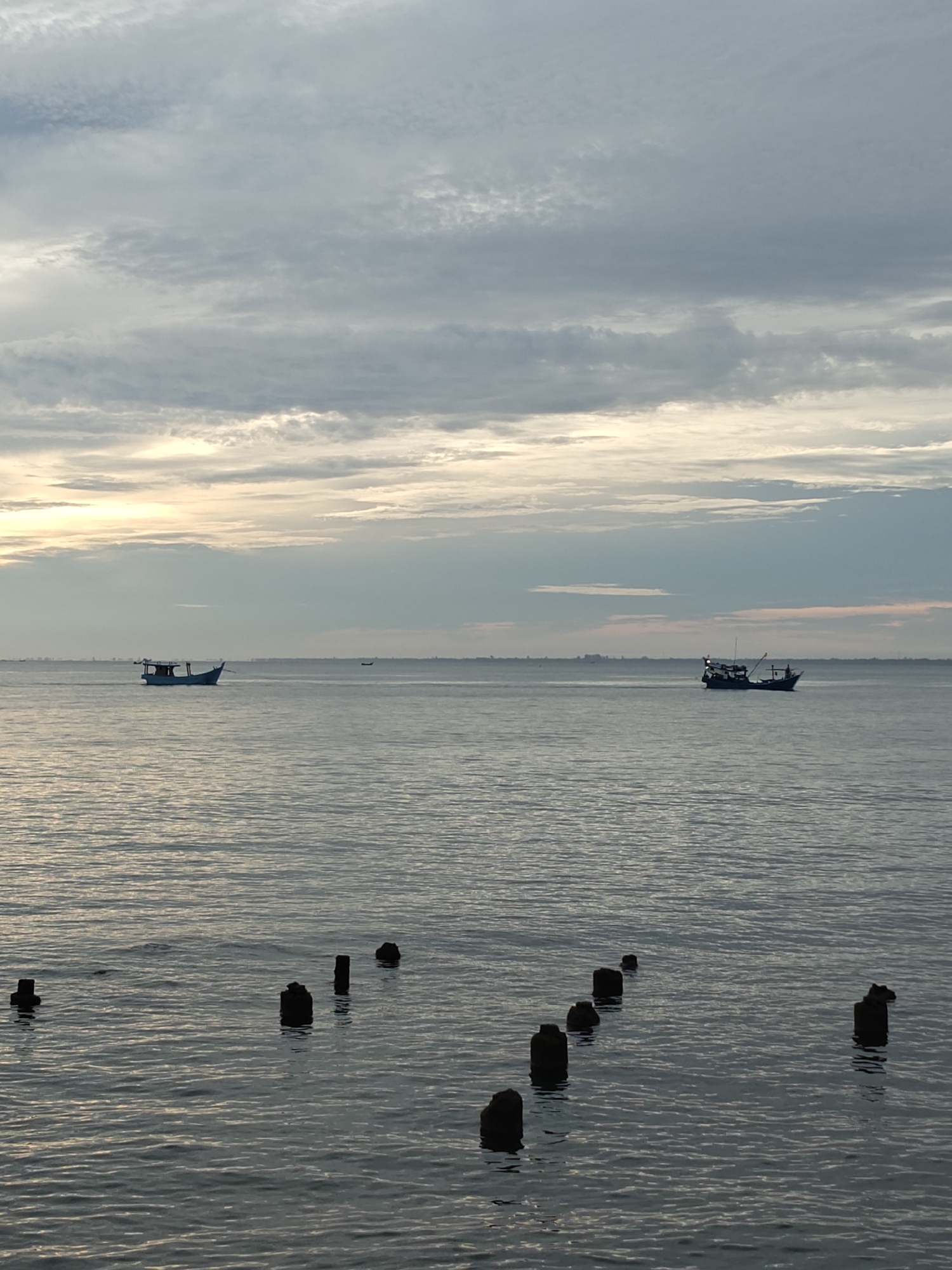 This image depicts a coastal seascape at twilight, likely in Lhokseumawe, Indonesia. The sky occupies the upper two-thirds of the frame, filled with stratocumulus clouds in shades of light grey, white, and muted blue. A subtle horizontal band of warm, yellowish light is visible along the lowest portion of the sky, indicating a low sun position. The horizon line reveals a distant, low-lying landmass or urban silhouette. The lower third of the image features the calm, rippled surface of the sea, reflecting the subdued light from the sky. In the foreground, seven dark, weathered pilings or posts are partially submerged in the water, extending diagonally from the lower left to the mid-right. Two fishing boats are present in the mid-ground. On the left, a light-colored boat, appearing blue and white, with an visible cabin structure, navigates or floats on the water. To the right, a second, darker-hulled fishing vessel, equipped with a mast and rigging, is also present. A third, smaller, dark silhouette of a boat is faintly discernible further in the distance between the two main vessels. No individuals are visible. The scene is illuminated by diffused natural light.