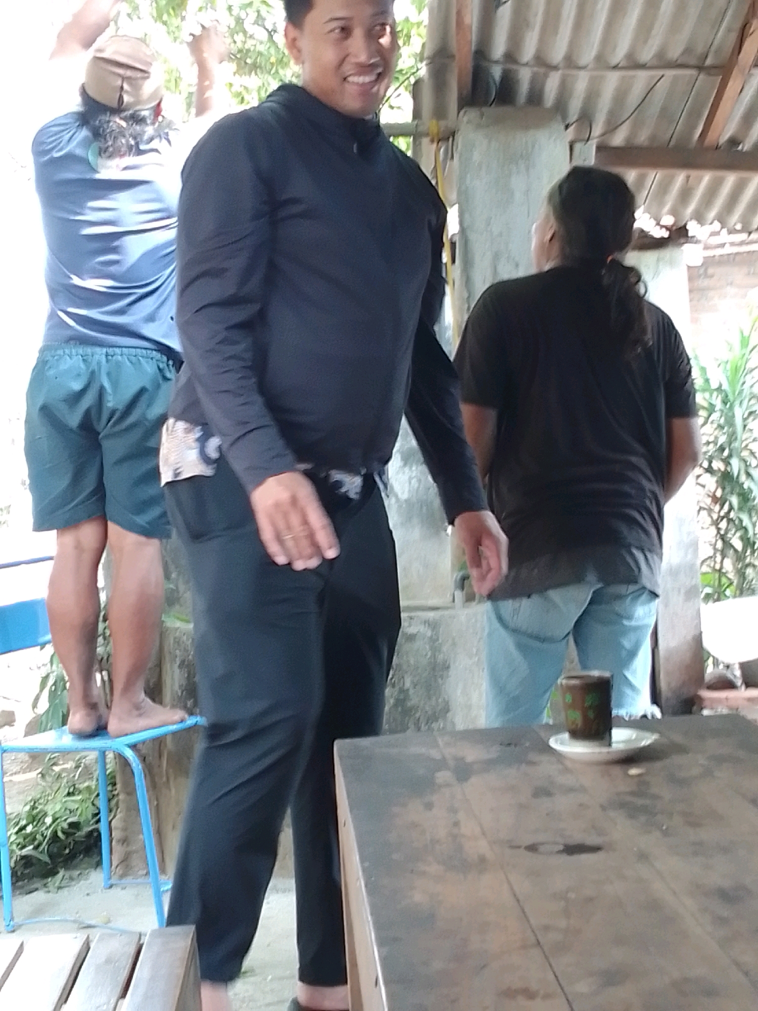 The image depicts an outdoor scene with three individuals. The primary subject, a man in the foreground, is wearing a black long-sleeved shirt and dark pants, smiling broadly. To his left, another person, partially obscured, wears a blue long-sleeved shirt and dark shorts, standing on a blue metal chair. To the right, a third individual, viewed from the back, wears a black t-shirt and light blue jeans. They are standing near a table where a small cup and saucer are placed. The environment includes a corrugated metal roof, concrete pillars, and lush green foliage. Based on the context, the scene is likely located in Trenggalek, Indonesia.