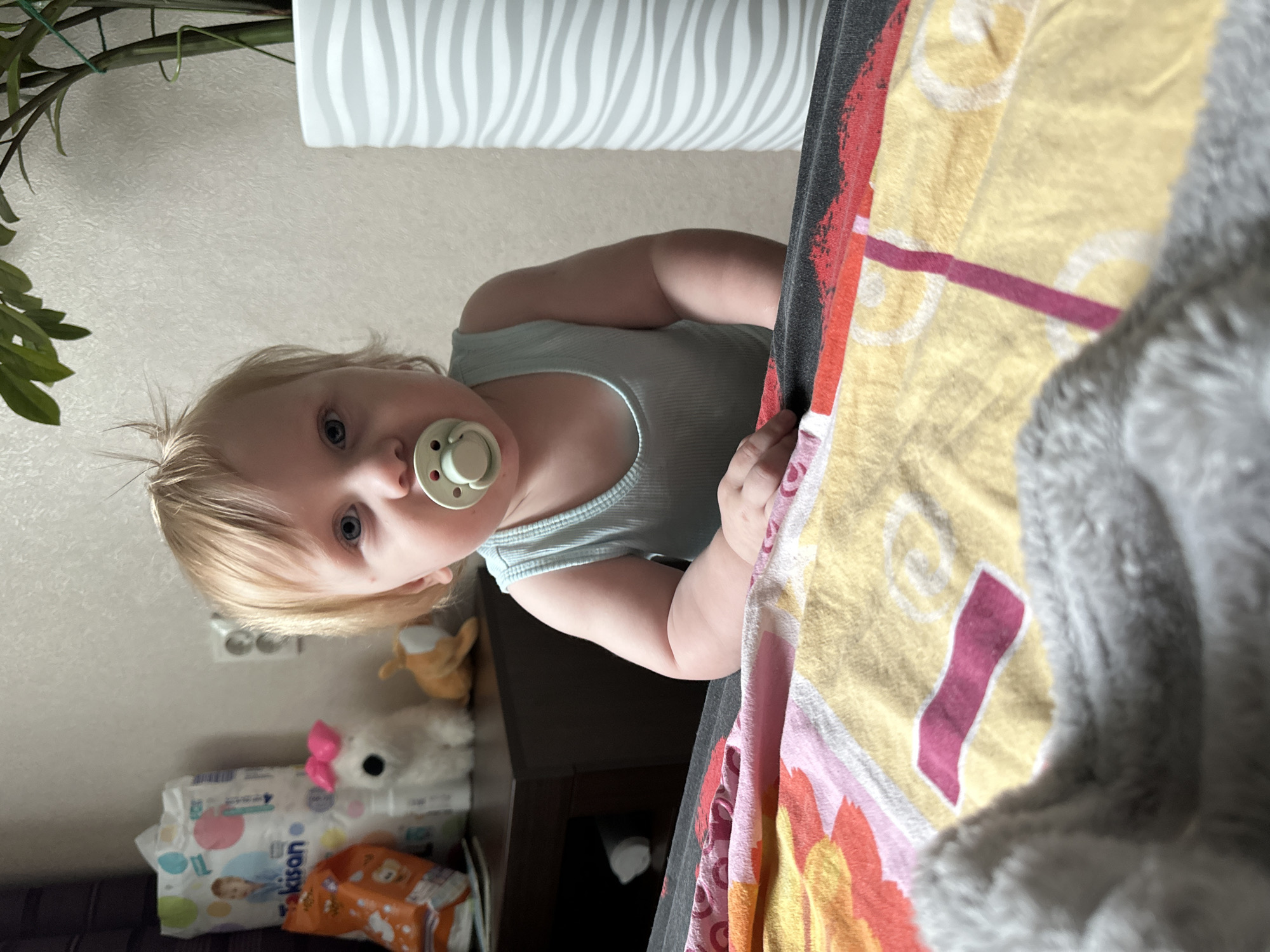 A toddler with blonde hair and blue eyes is sitting in front of a bed with colorful bedding. The toddler is wearing a light blue tank top and has a pacifier in their mouth. In the background, there is a shelf with various items, including a pack of diapers, a stuffed animal, and a plant in a striped vase. The room appears to be a bedroom. The image was taken in Sayanogorsk, Russia.