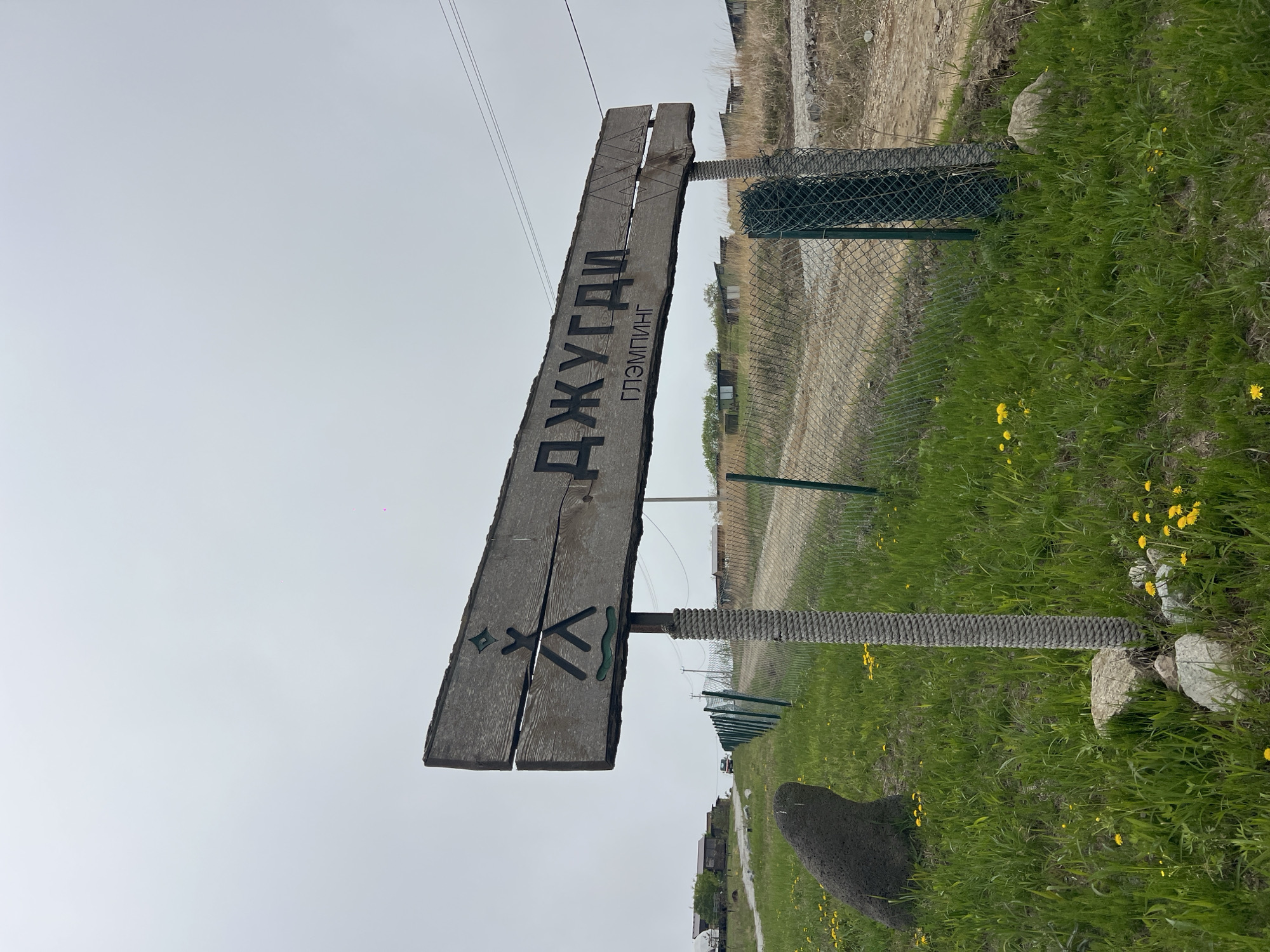 The image depicts a weathered wooden sign indicating "Джугди Глэмпинг" (Jugdi Glamping), likely an advertisement for a glamping site. The sign is made of two planks mounted on posts wrapped in rope. Below the text are symbols resembling a diamond shape, crossed sticks, and a wavy line. The sign stands beside a chain-link fence in a grassy area with yellow wildflowers. A dirt road runs parallel to the fence. In the background, there are buildings, possibly part of a village or campsite. The image suggests a rural setting, and the location is Zarubino, Russia. No people are visible.