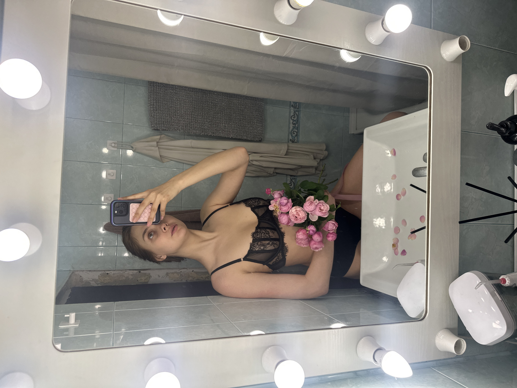 A young woman in a black lace bra and black shorts is lying on a tiled floor in what appears to be a bathroom. She is holding a bouquet of pink roses in one hand and a smartphone in the other, seemingly taking a selfie in a vanity mirror. The mirror is framed with light bulbs. The bathroom has tiled walls, a sink, and a towel rack with towels hanging on it. The sink contains scattered pink flower petals. The exact location in the city of Krasnoyarsk, Russia is not discernible from the image.
