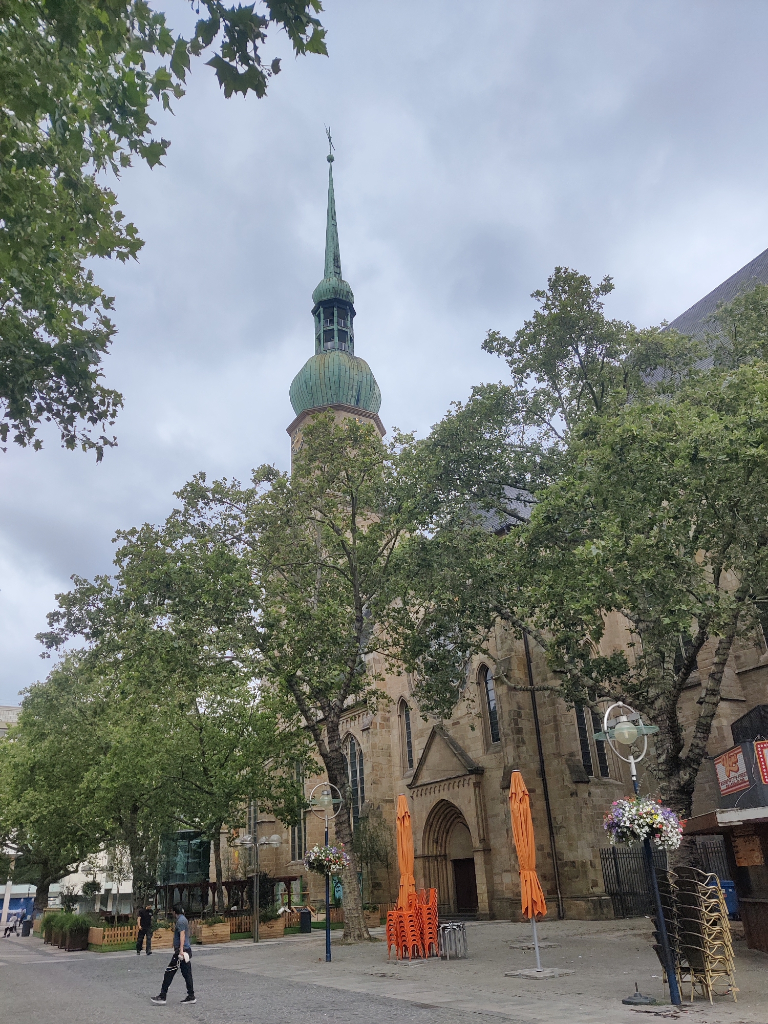 The image shows a view of the St. Reinoldi's Church in Dortmund, Germany, on a cloudy day. The church features a tall, green-domed spire with a cross at its apex. The facade of the church is made of light brown stone, with arched windows and entrances. In front of the church, there are several large trees with green leaves, and some are partially obscuring the building.
On the paved area in front of the church, there are orange outdoor umbrellas and stacks of chairs, suggesting an outdoor seating area for a cafe or restaurant. A street lamp with a flower basket hangs to the right, and stacks of chairs are also visible near it. Two men are walking on the paved area. One man in the foreground is wearing a grey t-shirt and black pants, carrying what appears to be a white bag. Another man in the background is wearing a black t-shirt and dark pants. In the distance, there are some buildings and planters with greenery. The sky is overcast, with grey clouds.