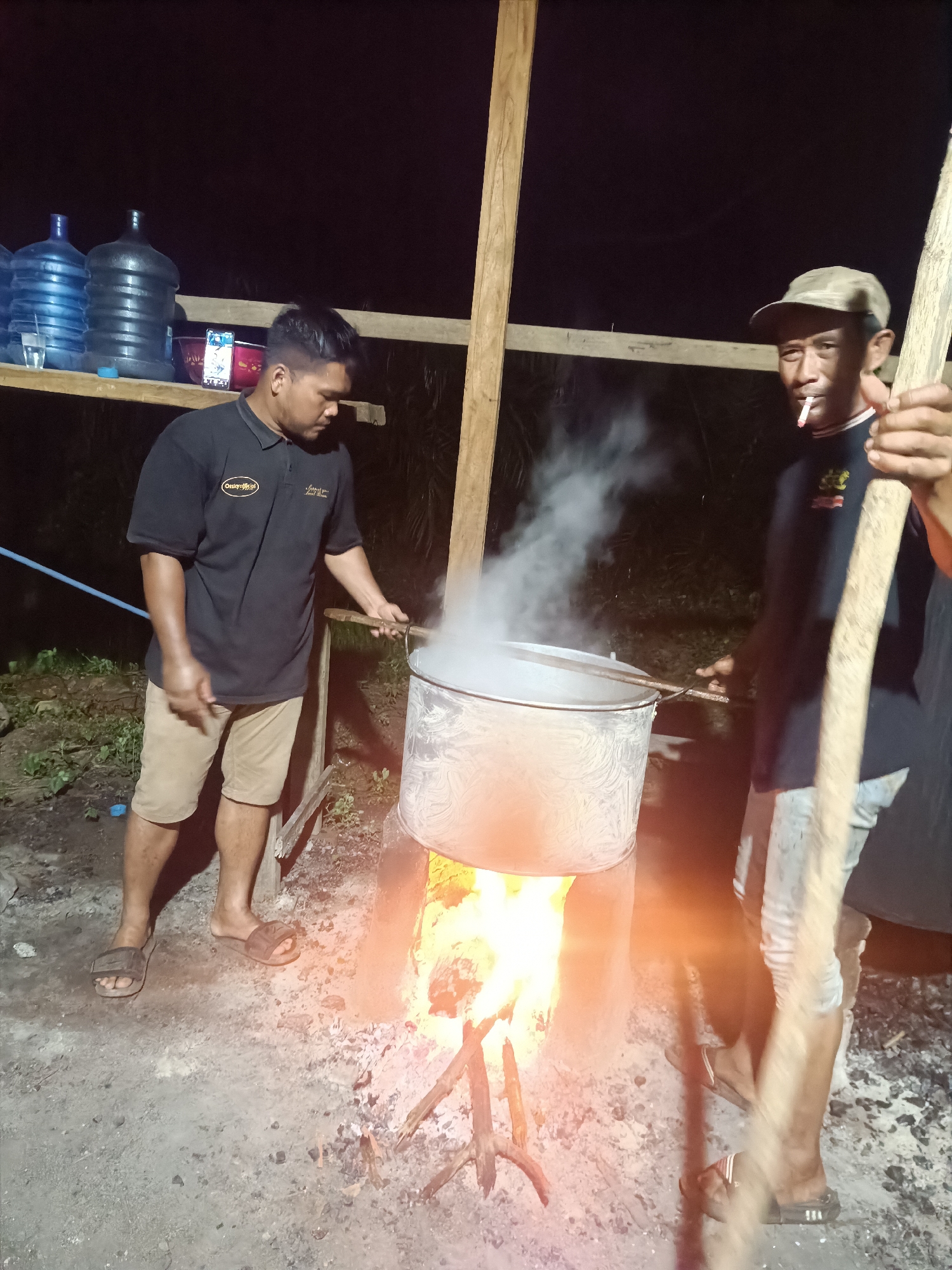 The image depicts an outdoor cooking scene at night, likely in Kilometer Delapan, Indonesia. Two men are attending to a large metal pot placed over an open wood fire, from which considerable steam is rising.
The man on the left, wearing a black polo shirt with a visible "Omiyorg official" logo, tan shorts, and dark sandals, is looking downwards, holding a stick or utensil towards the pot. He appears focused on the cooking process. The man on the right, wearing a light-colored cap, a dark t-shirt, light-colored pants, and dark sandals, holds a long wooden pole and has a lit cigarette in his mouth, looking towards the camera.
Behind the men, a rustic wooden structure serves as a makeshift shelf. On this shelf are several large plastic water dispensers, some full with blue-tinted water, along with a small glass, a red bowl, and a smartphone with an illuminated screen. A blue hose is visible on the ground near the man on the left.
The scene is illuminated primarily by the bright orange flames of the wood fire, casting a warm glow on the pot and the ground, contrasting with the dark background which indicates it is nighttime. Faint outlines of vegetation can be seen in the distance. The overall setting suggests a practical, possibly communal, outdoor cooking or boiling activity using traditional methods.