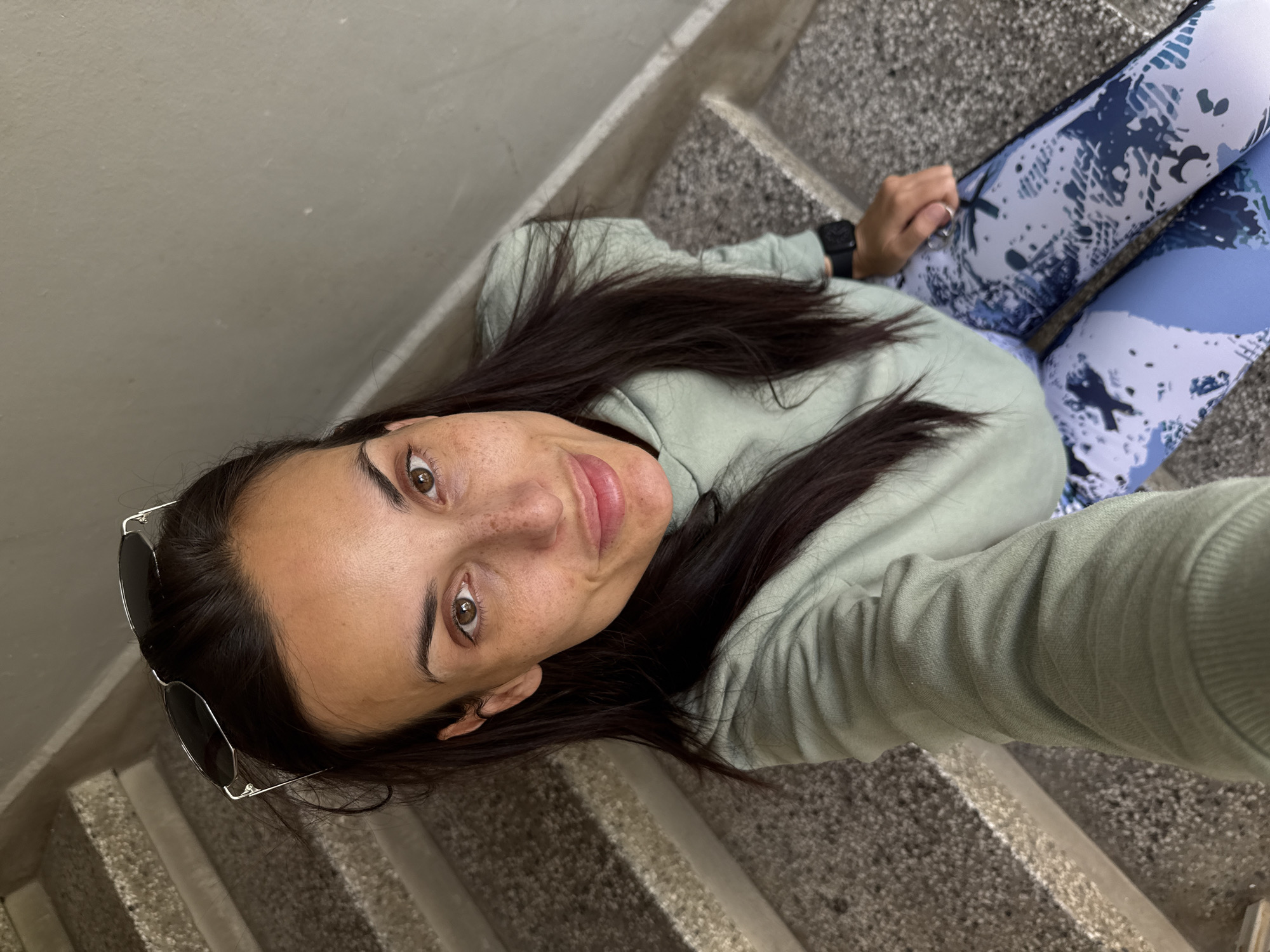 A woman is seated on gray concrete stairs, looking up at the camera. She has dark hair, brown eyes, and freckles. Sunglasses are tucked into the side of her hair. She wears a light green sweatshirt and blue patterned leggings. A black wristwatch is visible on her wrist. The background consists of the staircase and a light gray wall. The photo appears to have been taken in Pleven, Bulgaria.