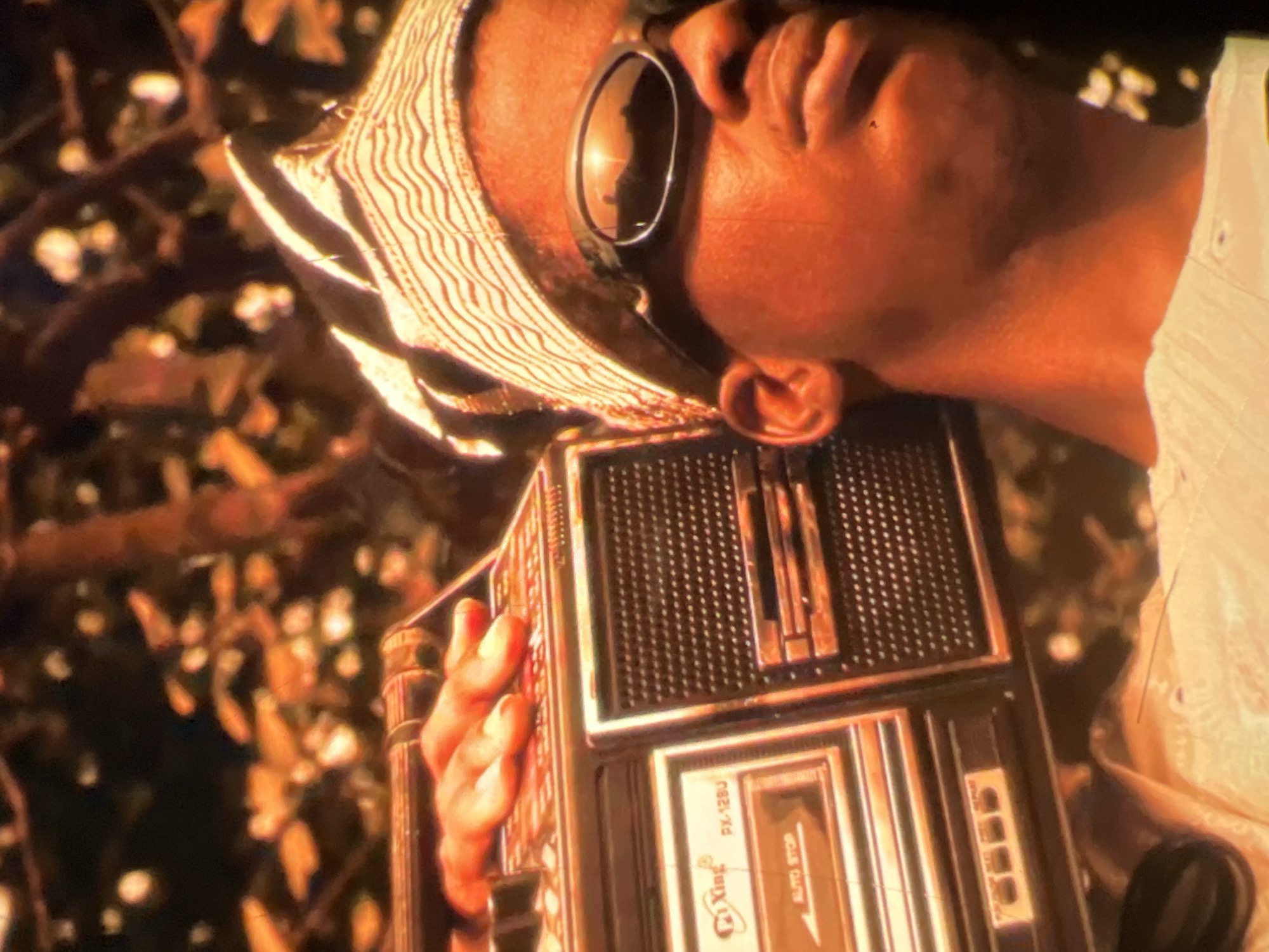 A close-up shot shows a person wearing a white dashiki and a patterned headwrap, with sunglasses on, and a boombox resting on their shoulder. The background features out-of-focus foliage, suggesting an outdoor setting. The lighting is warm and appears to be during sunset or golden hour. The boombox is a portable cassette player with visible buttons and speaker grills. The image is vibrant and possibly depicts a scene from Lagos, Nigeria, given the cultural attire.
