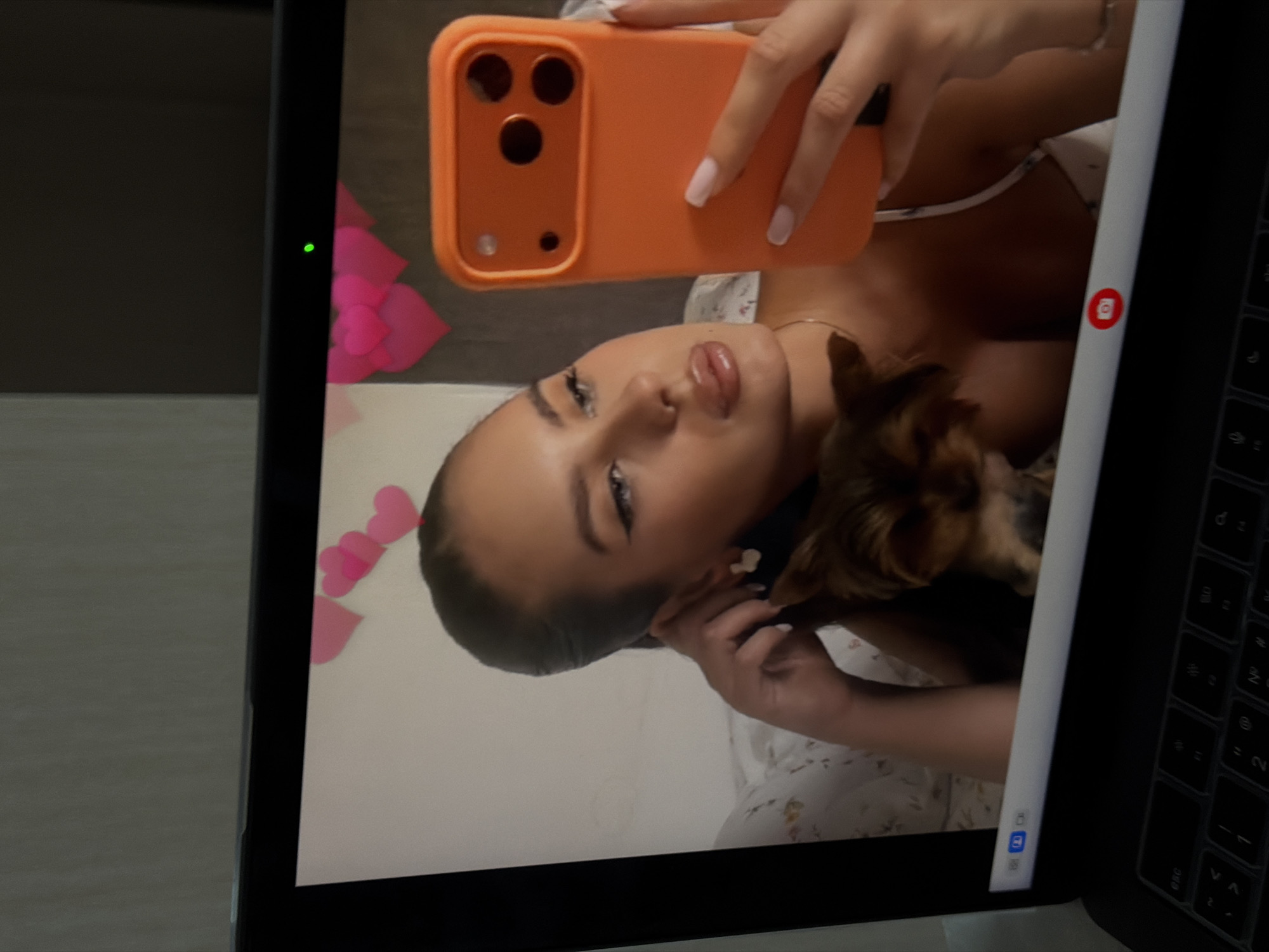 A close-up view of a laptop screen displaying a rotated image of a woman taking a selfie with an orange smartphone. A small brown dog is partially visible in the frame. The background appears to be a bed with a floral-patterned cover. Pink paper hearts are visible in the background of the screen. The laptop's keyboard is partially visible on the right side of the screen. The location of this scene in Krasnoyarsk, Russia, is not discernible from the image.