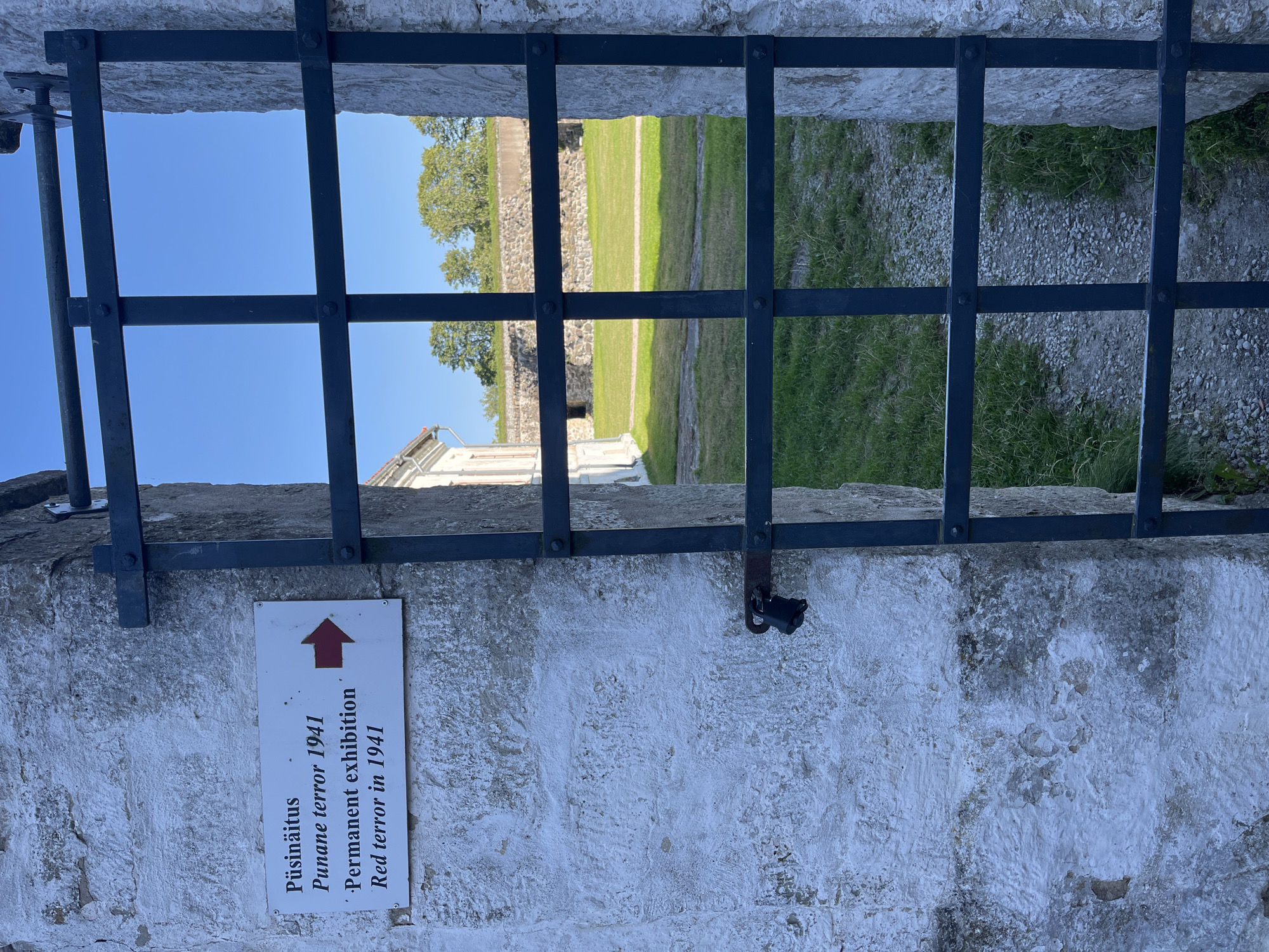 The image shows a white stone wall with a sign and a locked metal gate. The sign is rectangular and white with black text and a red arrow pointing to the right. The text on the sign reads: "Püsinäitus", "Punane terror 1941", "Permanent exhibition", and "Red terror in 1941". A black padlock is attached to the gate. Beyond the gate, there is a grassy courtyard with stone walls and trees in the background. The sky is clear and blue. The location is likely the Kuressaare Castle in Kuressaare, Estonia, as this is a known historical site that exhibits on the "Red Terror".