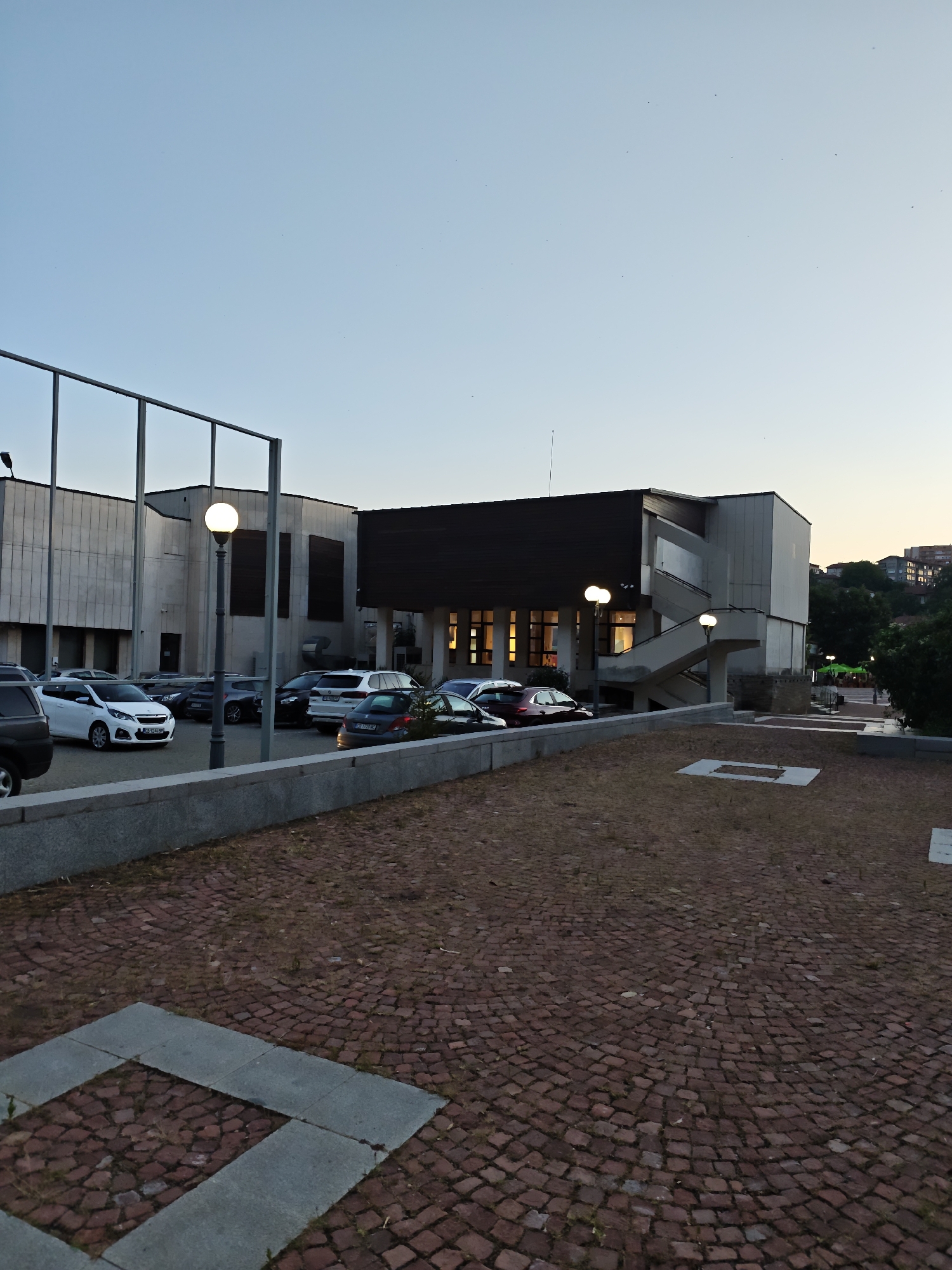 The image depicts an outdoor scene at dusk, featuring a modern building complex with a parking lot and a paved plaza in the foreground. The building is a multi-story structure with a contemporary architectural style, characterized by concrete and stone facades. A dark-colored, angled roof overhangs a section of the building, beneath which windows glow with warm interior light. An external staircase ascends along one side of the building. Several cars are parked in the lot, including a white hatchback, a dark SUV, and other various models. A tall streetlamp with a lit globe stands near the parking lot. The plaza in the foreground is paved with red cobblestones arranged in a circular pattern, with a rectangular inlay of larger gray stones. The sky is a pale, clear blue, transitioning to a lighter hue near the horizon, suggesting the end of daylight. The surrounding environment includes trees and buildings visible on a distant hillside. The scene is devoid of any visible individuals or overt actions. This location is identified as the area around the National Palace of Culture "Ukraina" in Lovech, Bulgaria.