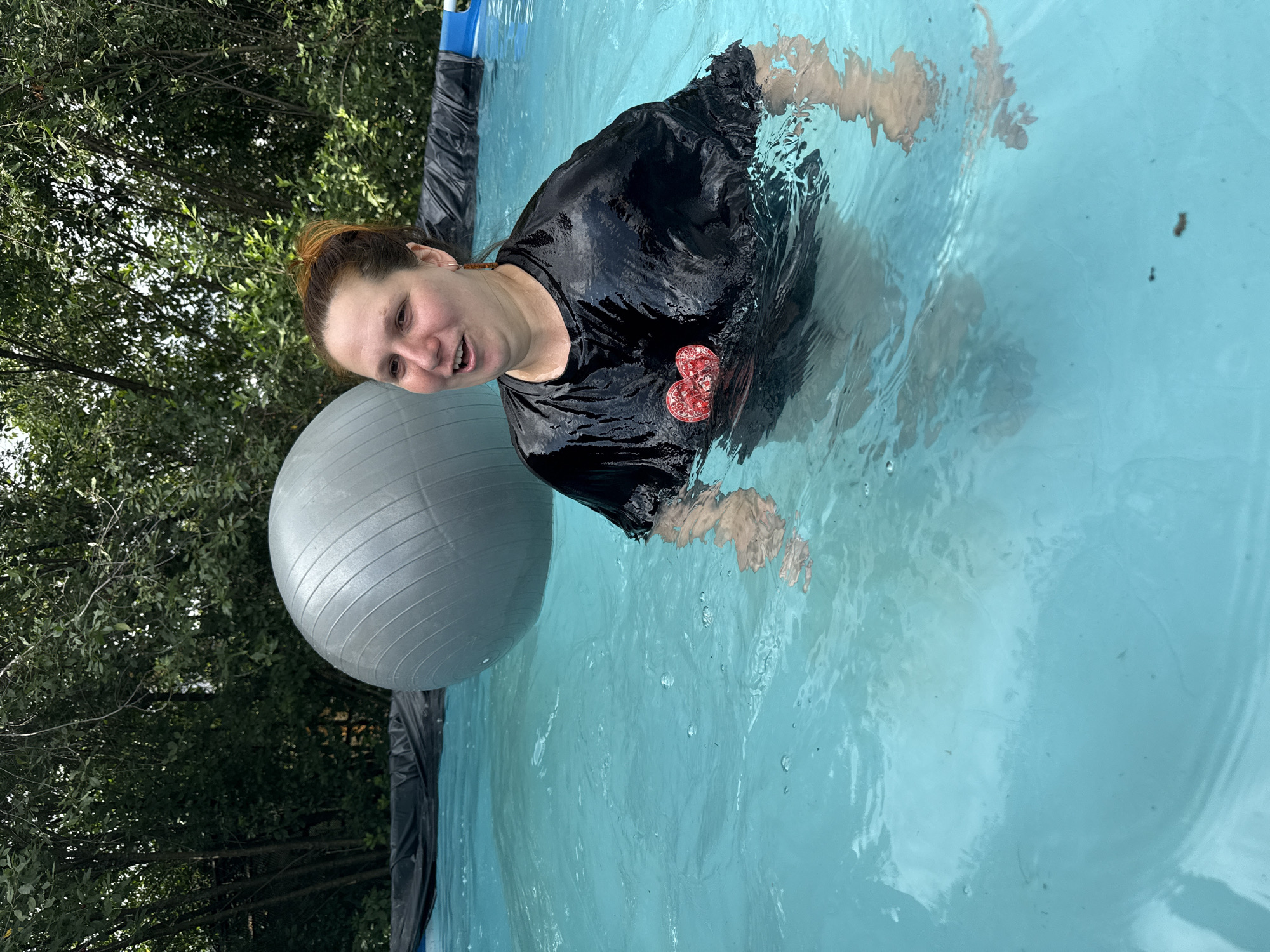 A woman with red hair pulled back in a bun is standing in a bright blue swimming pool. She is wearing a black t-shirt with a heart-shaped emblem on the chest. Her t-shirt is wet and clinging to her. Behind her, a large grey exercise ball bobs in the water. Trees and foliage line the edge of the pool, suggesting an outdoor setting. The scene appears to be in the city of Khokhryaki, Russia.