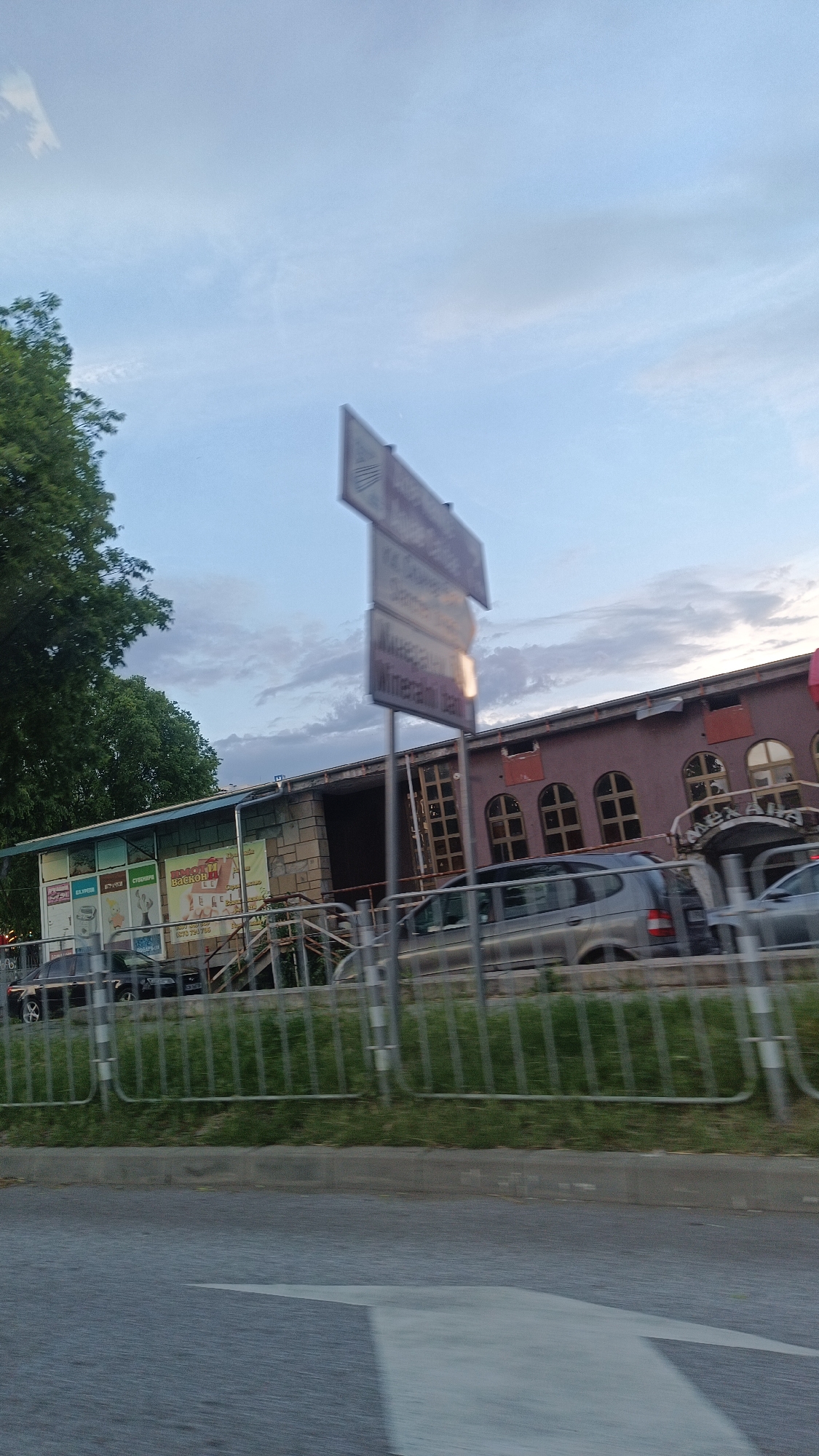 The image shows a street scene in Burgas, Bulgaria, taken from a moving vehicle. A blurred white directional arrow is visible on the road surface in the foreground. A metal fence with vertical bars separates the road from the sidewalk. Behind the fence, several cars are parked in front of a complex of buildings.
The buildings consist of two distinct sections. The left section appears to be a one-story structure with a blue and white awning, covered in advertisements. The right section is a two-story building with a stone facade section. It has arched windows on the upper level. The building on the right has an entrance way that looks like it reads "Mexana".
A tall, blurred street sign stands between the buildings. In the background, the sky is a mix of blue and cloudy skies. A tree is visible on the left side of the frame.