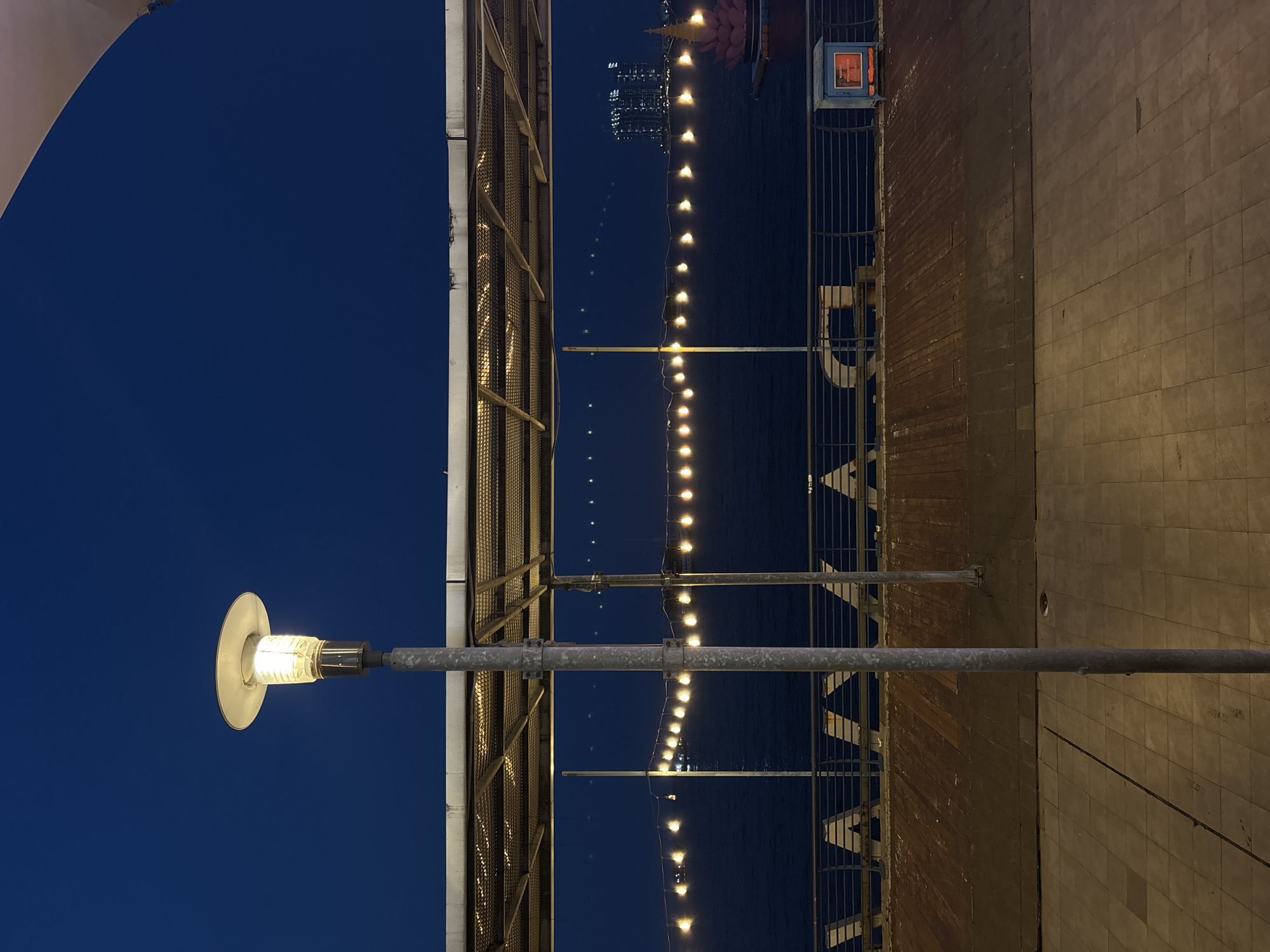 Captured from a low-angle perspective, the image presents a nighttime urban waterfront scene. A street lamp glows brightly in the lower left corner, casting light onto a paved walkway lined with a metal railing. A string of decorative lights stretches across the water, reflecting on the dark surface. In the distance, city lights are visible. The scene is in the city of North Jakarta, Indonesia.
