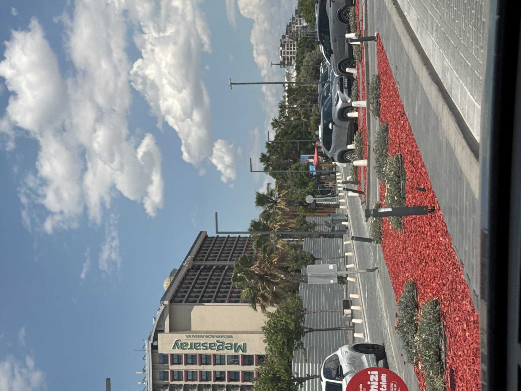 The image shows an outdoor scene in Konyaaltı, Turkey, with a row of parked cars along a curb. A strip of red decorative ground cover separates the parking area from the sidewalk. The Megasaray hotel building is visible in the background, with palm trees planted in front. The sky is blue with white clouds. Streetlights are present along the road. A white car is visible in the foreground.