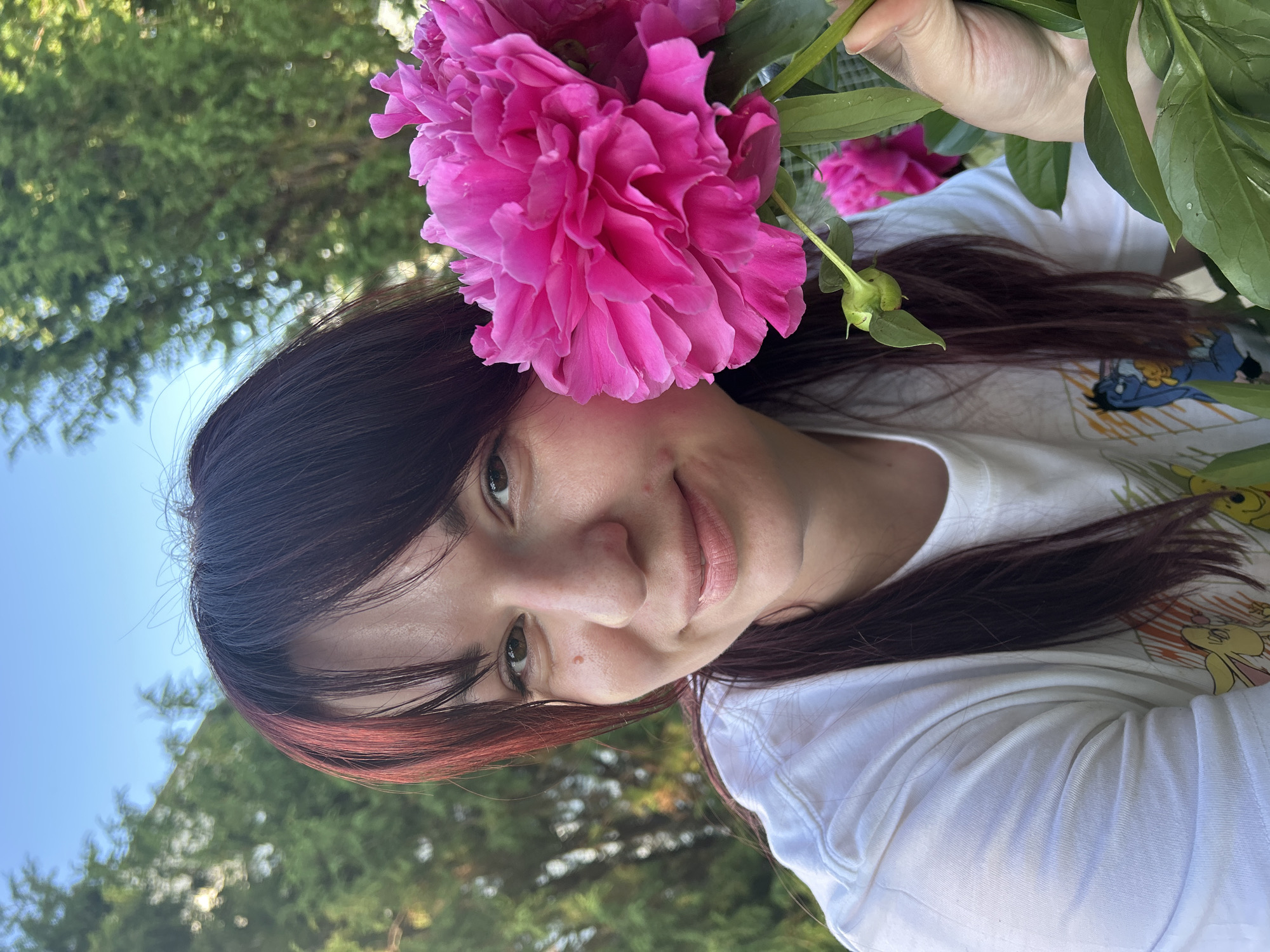 A woman with dark, reddish-brown hair and bangs is shown in a close-up, outdoor portrait, holding a bright pink peony flower. She is wearing a white t-shirt with a cartoon graphic. The background consists of green foliage and a blue sky, suggesting a natural, possibly garden or park setting. The image appears to have been taken during daylight hours. No specific location in the city of Фоки, Russia is visible or indicated in the image.
