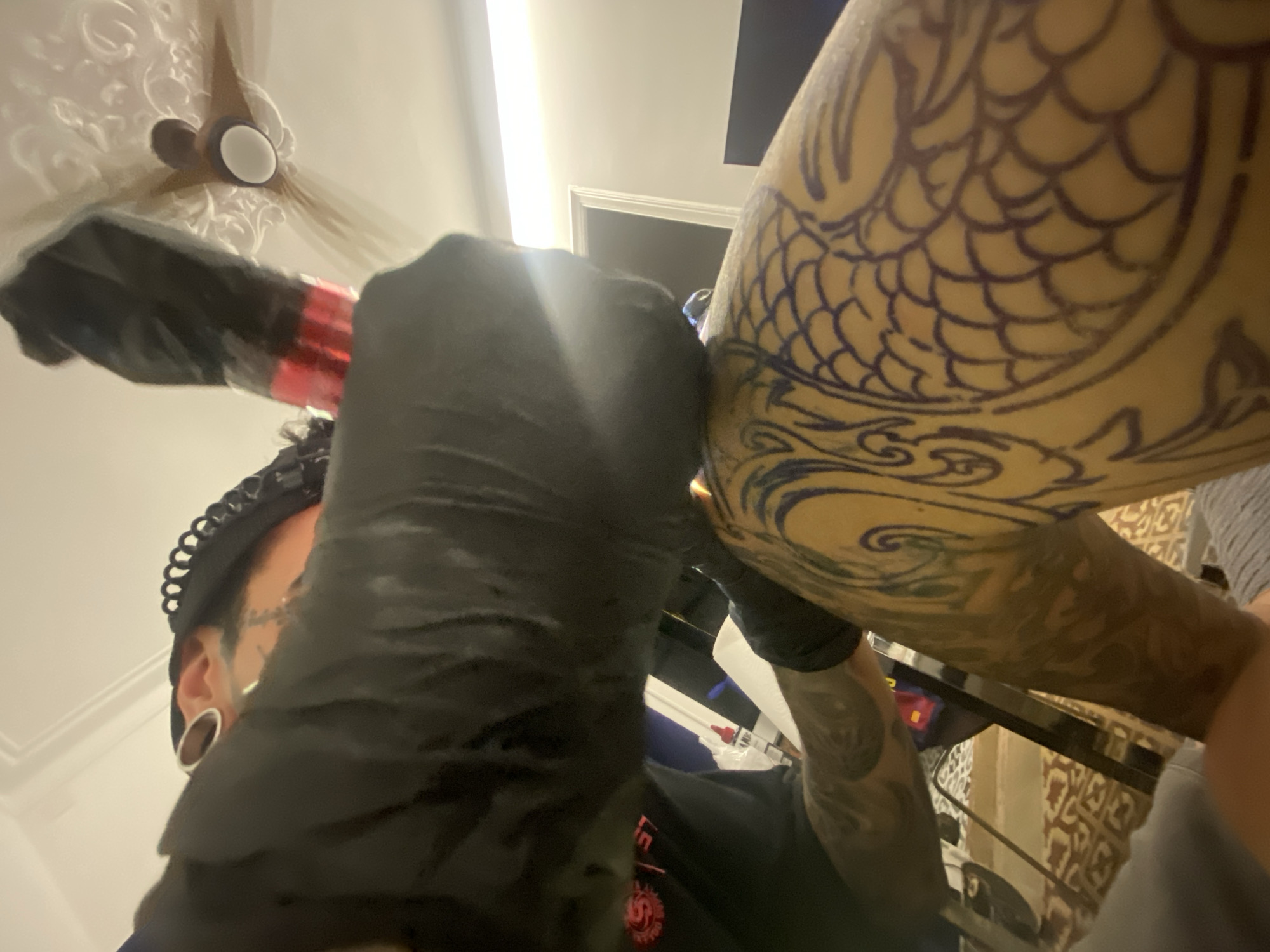 A tattoo artist is captured in a low-angle shot as they are working on a large tattoo on a person's arm. The tattoo appears to be a koi fish design. The artist is wearing black gloves and a black cap. There is visible tattooing on the artist's neck and arms. There is tattoo equipment (including a tattoo machine) visible in the artist's gloved hands. The setting seems to be indoors, possibly in a tattoo studio. A ceiling fan can be seen in the upper part of the image. The location appears to be Barcelona, Spain, based on contextual evidence like the ceiling fan and the artistic style that may be indicative of the region.