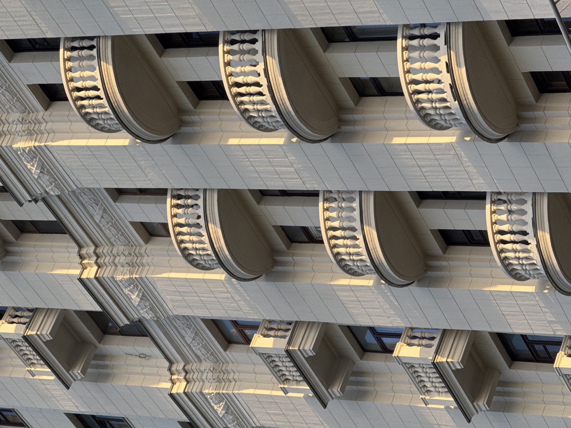 The image shows the facade of a light-colored, ornate residential building, likely in Moscow, Russia, illuminated by the warm light of the setting sun. Several balconies with white balustrades and decorative elements are visible, protruding from the building. The balconies are of varying sizes and shapes, some appearing rounded and others more rectangular. Rows of windows with dark frames are also present, recessed within the facade. The architectural style suggests a pre-war Stalinist or neoclassical design, common in older Moscow districts. The building's exterior is constructed with stone or a similar material, with decorative molding and detailing visible along the edges of the balconies and around the windows. The lighting creates strong shadows that highlight the three-dimensional aspects of the architecture, emphasizing the texture and depth of the facade. No people are visible in the image. The overall impression is one of grand and historic urban architecture.