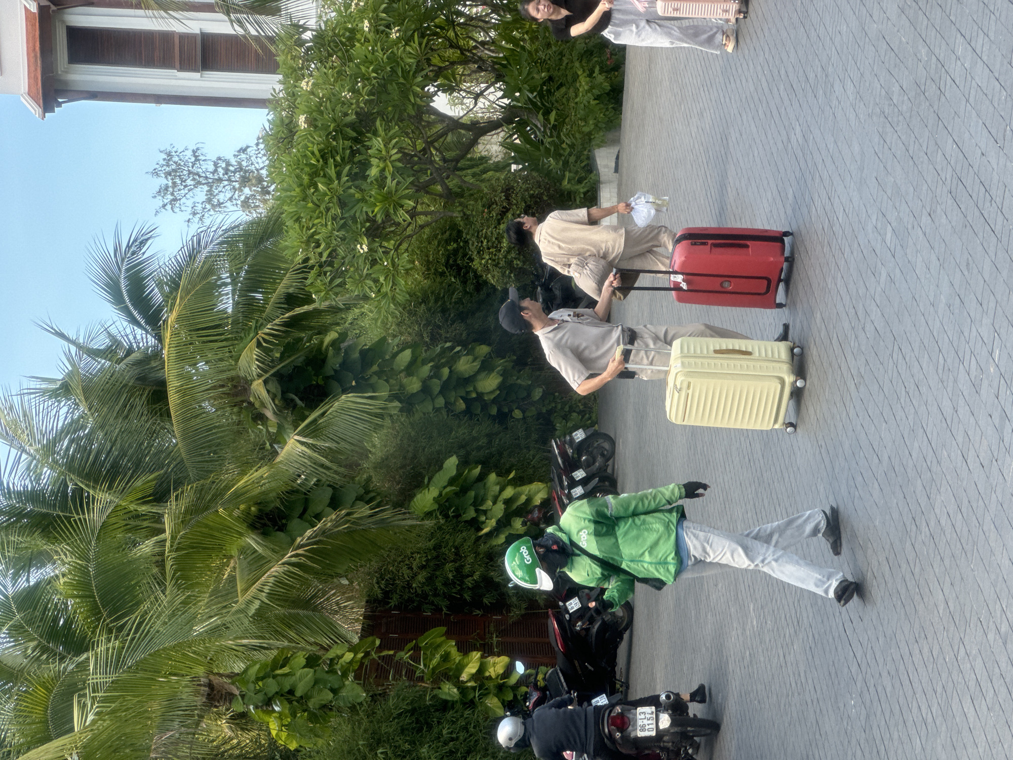 The image shows a street scene in Phu Thuy Ward, Vietnam. A man in a light grey uniform and baseball cap pulls a yellow suitcase, followed by a man in beige clothing pulling a red suitcase. In the foreground, a delivery driver in a green jacket and helmet stands beside several parked motorbikes. Other people are walking in the background. Lush tropical foliage includes palm trees. The ground is paved. A building is partially visible at the top left of the image.