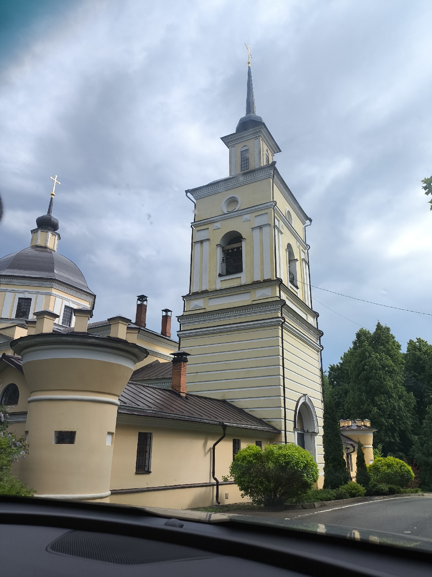 The image shows a yellow and white Orthodox church complex under a cloudy sky. The complex includes a tower with a tall, pointed spire and a dome-shaped structure topped with a cross. Smaller buildings and landscaping are visible at the base of the structures. The photograph was taken from inside a vehicle, with the dashboard visible at the bottom of the frame. The location is unknown.