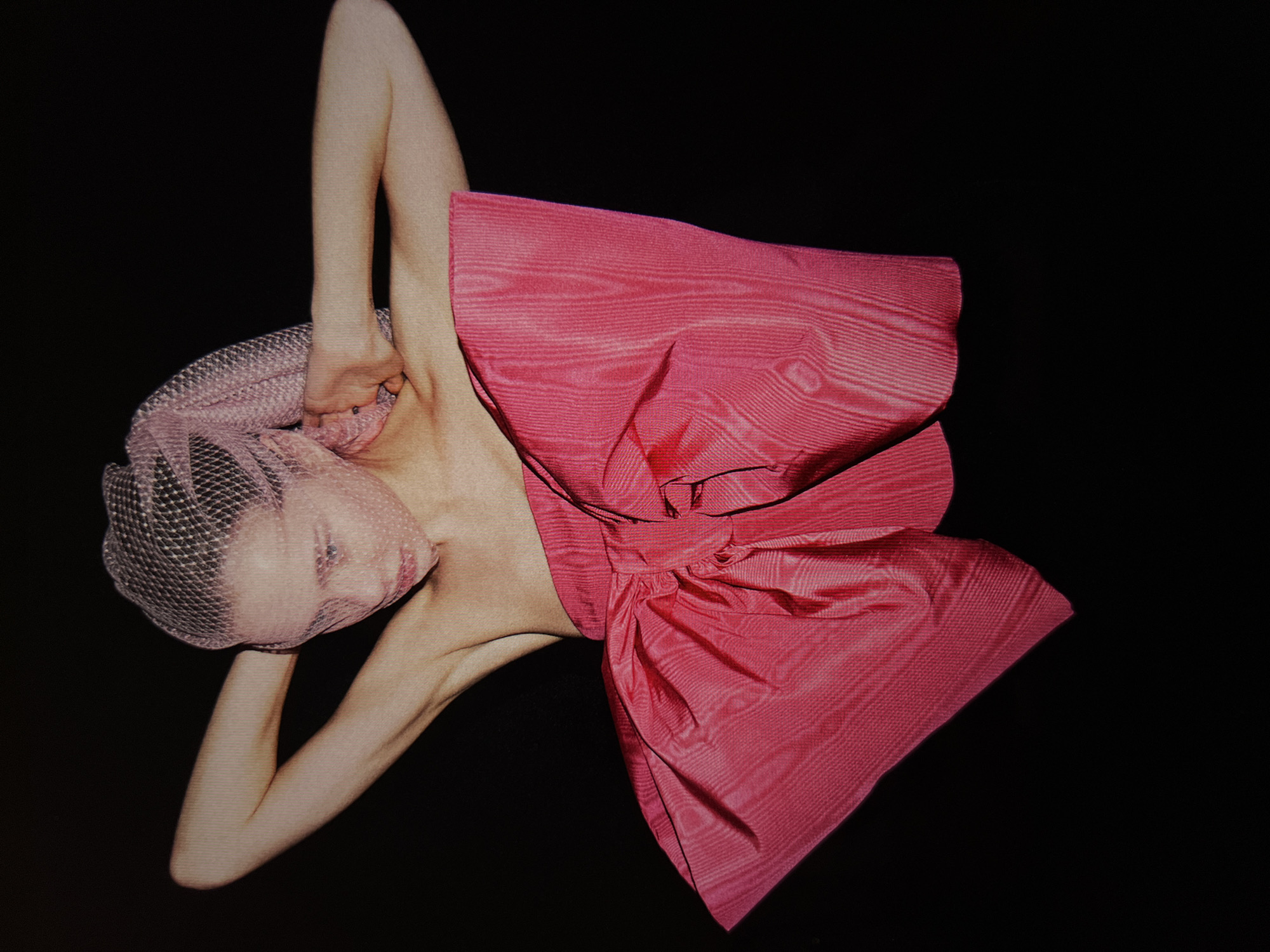 A person is seen against a black background. The person's head is covered by a pink mesh veil, and they are wearing a large, bright pink bow around their torso. Their arms are raised behind their head, with their hands touching their neck. The bow appears to be made of a textured fabric, possibly silk or satin, with a visible sheen. The lighting emphasizes the contours of the person's body and the folds of the bow.
