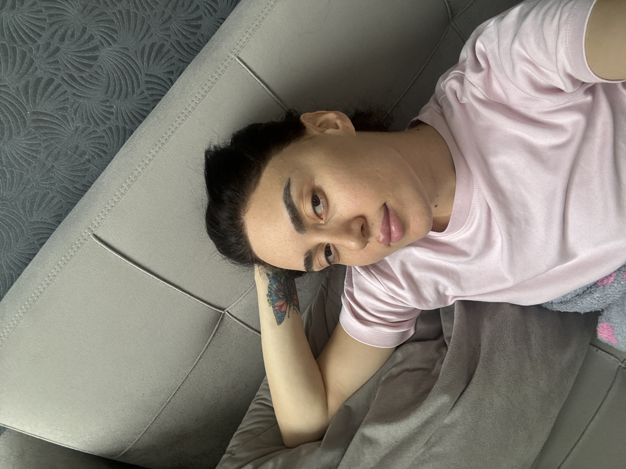 The image captures a person lying on a light gray upholstered sofa in Tbilisi, Georgia, looking directly towards the camera. The individual, appearing to be female, has dark hair pulled back and prominent, neatly groomed eyebrows. They possess a light complexion and are dressed in a plain, light pink short-sleeved t-shirt. A vibrant butterfly tattoo, rendered in shades of red, blue, and black, is visible on their right forearm, positioned on the sofa near their head.
A plain grey blanket partially covers the person and the sofa. To the right, a fragment of another grey fabric with pink patterns is visible, possibly a different blanket or clothing item. The sofa itself features distinct stitched seams and geometric paneling, creating a quilted effect. The background reveals a dark grey wall adorned with a textured wallpaper, showcasing a repeating, intricate pattern of fan-like or swirling motifs. The lighting is soft and even, suggesting an indoor setting. The person's expression is neutral and calm, with no visible text in the frame.