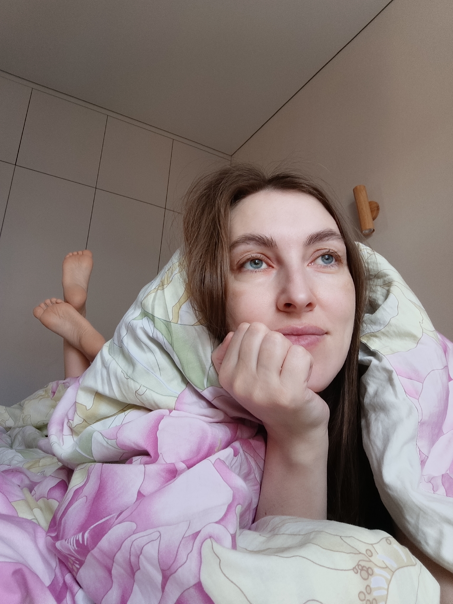 A woman is lying in a bed, partially covered by a floral-patterned blanket. She is resting her face on her hand and looking upwards with her blue eyes. Her bare feet are visible, raised in the air. A wooden fixture is mounted on the wall behind her, and a tiled wall is also visible. The photograph was taken in the city of Chaikovsky, Russia.