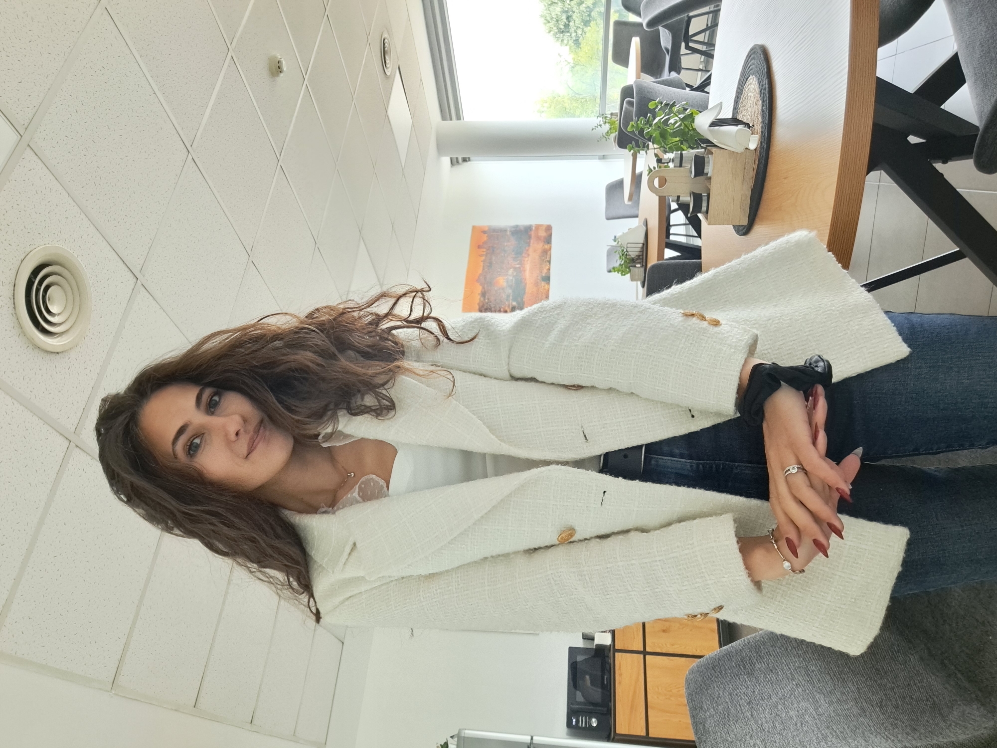 A woman is photographed indoors in Sofia, Bulgaria. She has long brown curly hair and is wearing a white tweed blazer over a white top and blue jeans. Her hands are clasped together and adorned with rings and a bracelet. The background includes office furniture, a table with a decorative arrangement, and a painting on the wall. The room has a white tiled ceiling with circular ventilation.