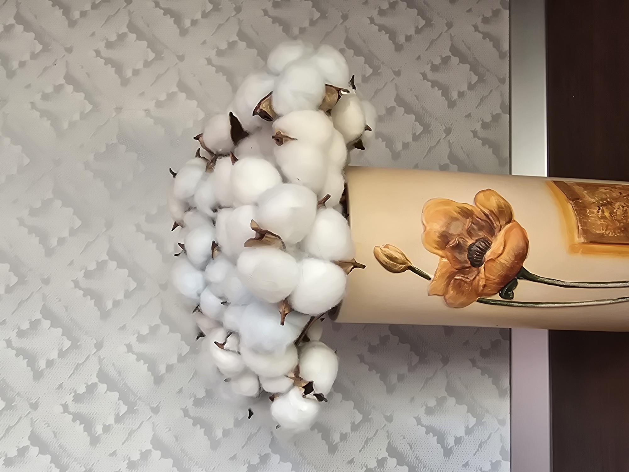 A vase filled with cotton bolls is displayed in a room in Sofiivska Borshchahivka, Ukraine. The vase is cylindrical and light beige with a painted orange poppy flower and a bud on its surface. The cotton bolls, which are fluffy and white with brown stems, overflow from the top of the vase. The background is a light gray wallpaper with a patterned texture. A dark brown surface, possibly a table, is visible at the bottom of the image.
