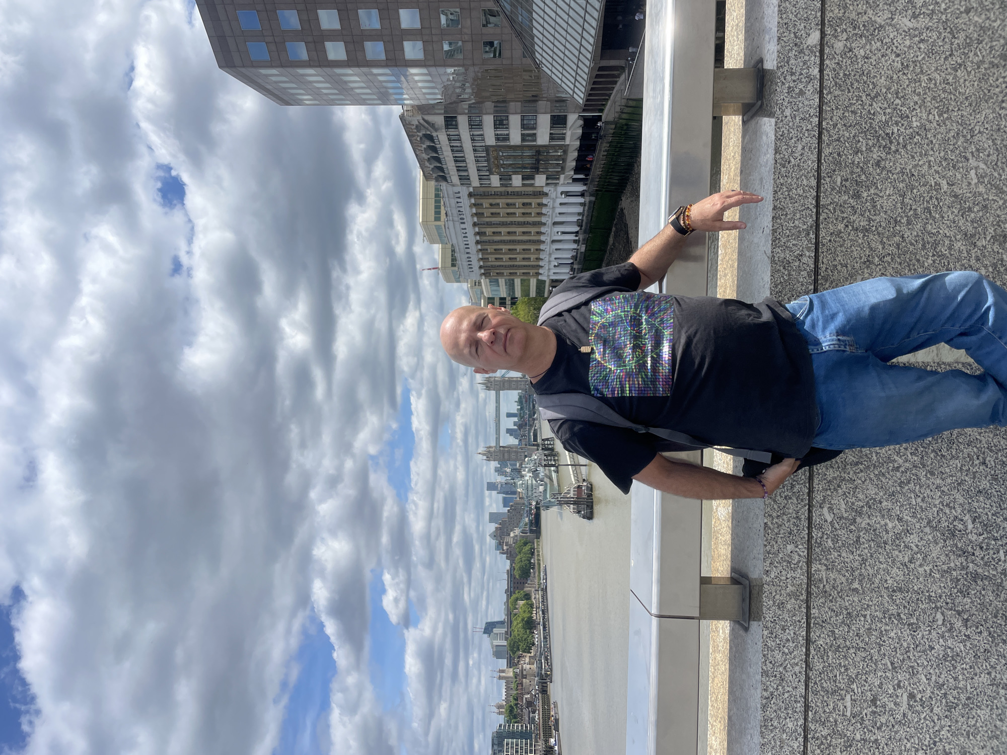 A bald, light-skinned man stands on a bridge overlooking the River Thames in London. He is wearing a black t-shirt with a colorful, geometric pattern and blue jeans. He has a backpack on and is leaning on the railing of the bridge. In the background, the Tower Bridge and other London landmarks are visible. The sky is cloudy with patches of blue.