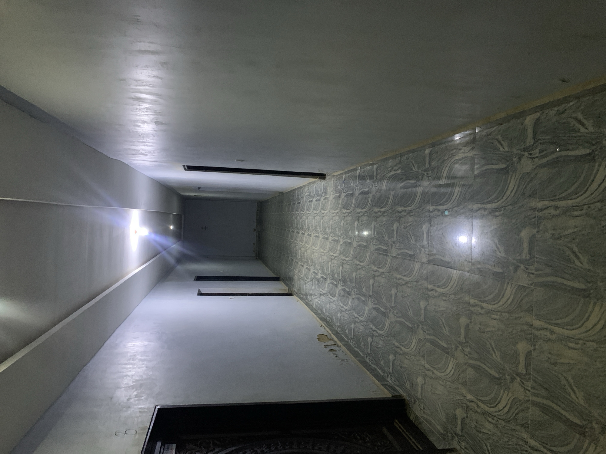 The image depicts an indoor hallway or corridor with a low-angle perspective. The left wall is a plain light gray, while the right wall is tiled with a gray and white marble-like pattern. The ceiling is plain white. There are several dark, horizontal recessed structures visible along the ceiling. At the far end of the hallway, a door is visible. The floor and walls appear to be reflective, possibly due to lighting. The floor is in front. The location is Kabong, Nigeria.