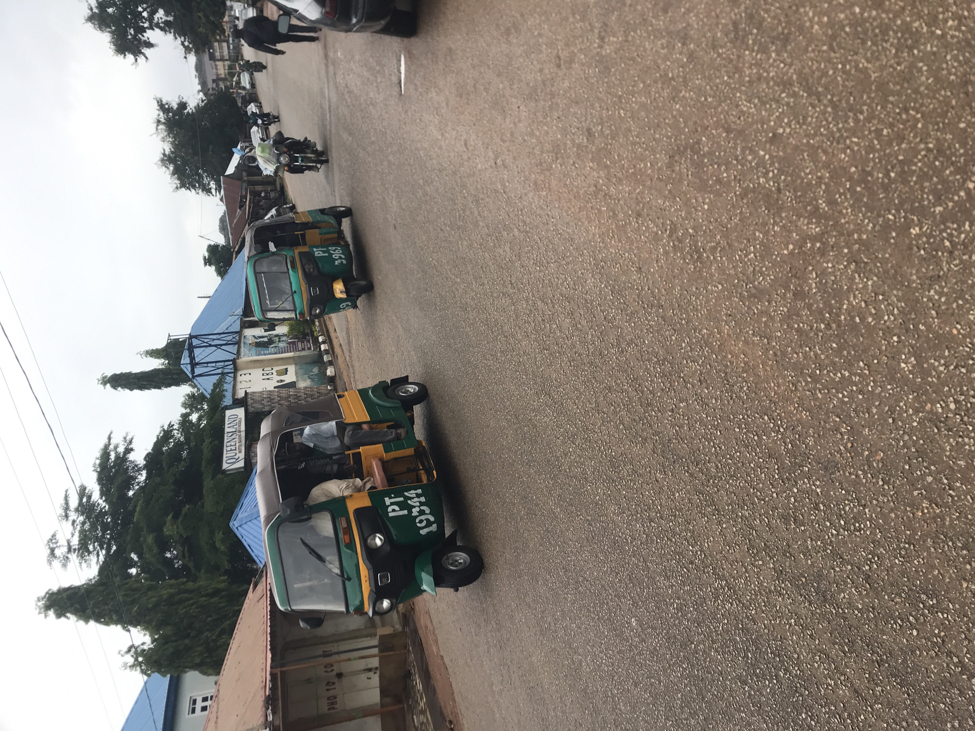 The image depicts a street scene in New Stadium, Nigeria, under an overcast sky. In the foreground, a green and yellow auto-rickshaw, identified by the license plate "PT 19344", is moving away from the viewer. The rickshaw has two passengers visible inside. To the left of this rickshaw is another similar vehicle, painted green and yellow, with the license plate "PT 3969". This second auto-rickshaw is moving towards the viewer and also has passengers.
Further down the street, several motorcycles are visible. One motorcycle, ridden by a person wearing a dark jacket, is moving away from the viewer and is laden with bags. Another person in a dark jacket is walking away from the viewer on the right side of the road. A black car is partially visible on the far right of the frame.
In the background, buildings line the street. On the left, a building with a brown, corrugated roof and a white section with windows is visible. A sign with the words "QUEENSLAND" is mounted on a building further back. Trees, including tall, slender ones and broader, leafy ones, are scattered throughout the scene. Electrical wires crisscross above the street. The road surface is asphalt, appearing wet in some areas. The overall impression is a moderately busy street with local transportation and commercial establishments.