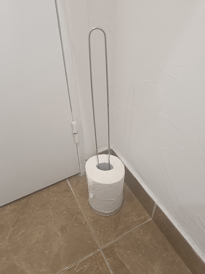 A toilet paper holder stands on a tiled floor beside a white door and wall. The holder is a simple, silver-colored frame with a tall, looped handle. The toilet paper is stacked high, nearly reaching the handle's base. The tiles are a warm, brown color with subtle veining. The wall has a slightly textured, painted finish. The overall scene is a clean, functional bathroom space.