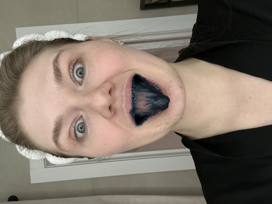 A woman poses with her mouth wide open, revealing a starkly blue-stained tongue. She has light skin, blue eyes, and wears a white headband. The background includes a tiled wall and a dark garment on her shoulder. Her expression is playful, suggesting she intentionally dyed her tongue. The focus is on the contrast between her natural features and the unusual, vividly colored tongue.