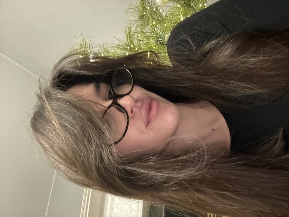 A female individual is depicted in an indoor setting, identified as the city of Rasskazovo, Russia. The subject occupies the majority of the frame, facing slightly downwards and to the viewer's right. She has long, layered brown hair with bangs that partially obscure her forehead. She is wearing dark, tortoise-shell rimmed eyeglasses. Her lips are slightly parted. A small, dark mole is visible on her upper neck, below her jawline, on the viewer's left side. She is attired in a dark, possibly black, crew-neck garment. In the background to the left, a light-colored wall with white molding is partially visible. To the right of the subject, a blurred green object, consistent with a pine or fir branch, is present, decorated with numerous small, illuminated white lights and at least one spherical white ornament. The lighting suggests an indoor environment.