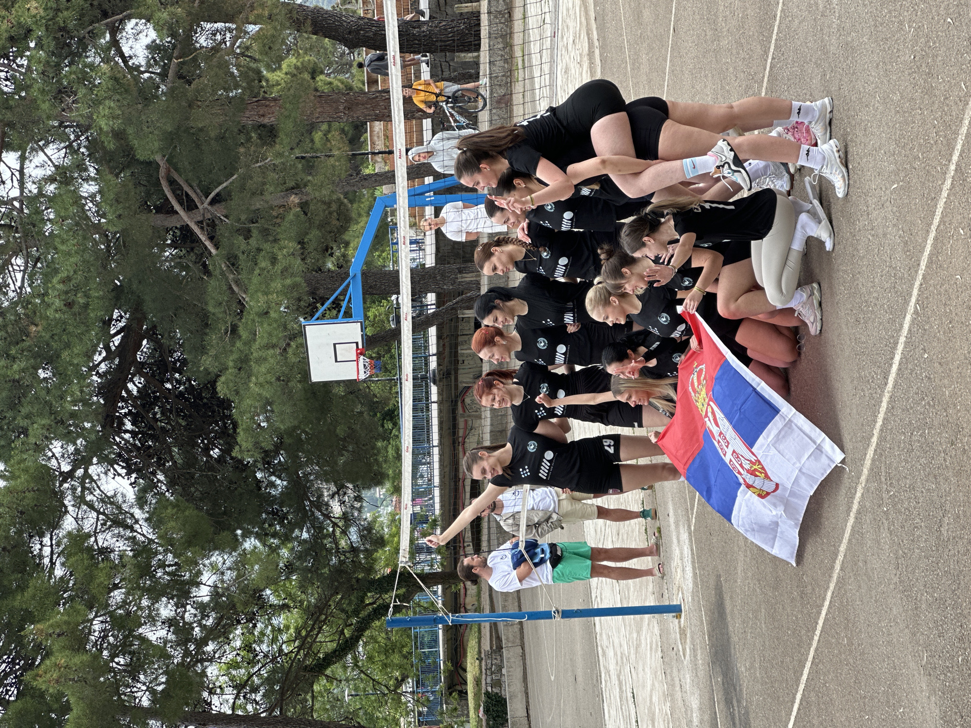 A group of 10 women are posed in a line on a paved surface, likely a basketball court or similar outdoor recreation area, under a light overcast sky in the city of Budva, Montenegro. Most of the women are wearing matching black tops with what appears to be a logo on the front, black shorts, and athletic shoes. They are arranged in a line, each partially overlapping the one in front, with the last few women on their hands and knees supporting the weight of the women behind them. The group is holding a flag of Serbia. Two men stand on the side, one of whom is barefoot and wearing green shorts.
Behind the group is a volleyball net strung between blue poles, a basketball hoop, and several trees. In the background, there's a glimpse of other people, including one person riding a bicycle. The overall setting suggests an outdoor sporting event or team celebration.