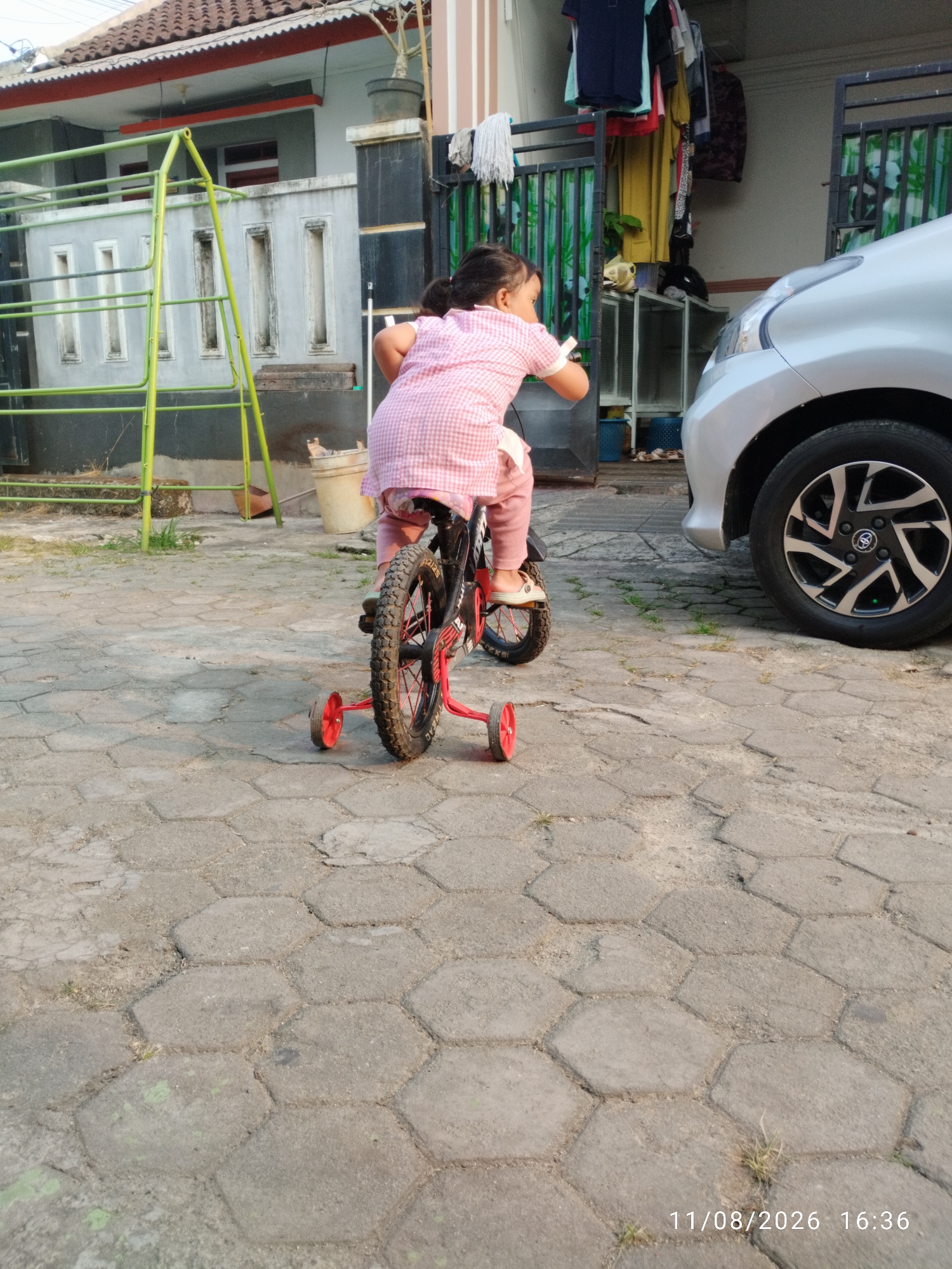 A young child, possibly a girl, is riding a small red bicycle with training wheels on a paved area. The child is wearing a pink checkered shirt and pink pants. The bicycle has black tires with red rims. In the background, there is a house with a red-tiled roof and a green metal playground structure. A silver car is parked to the right of the frame. The ground is covered in hexagonal paving stones. The timestamp in the lower right corner indicates the image was taken on August 11, 2026, at 16:36. The scene appears to be a residential outdoor space in Bandar Lampung, Indonesia.