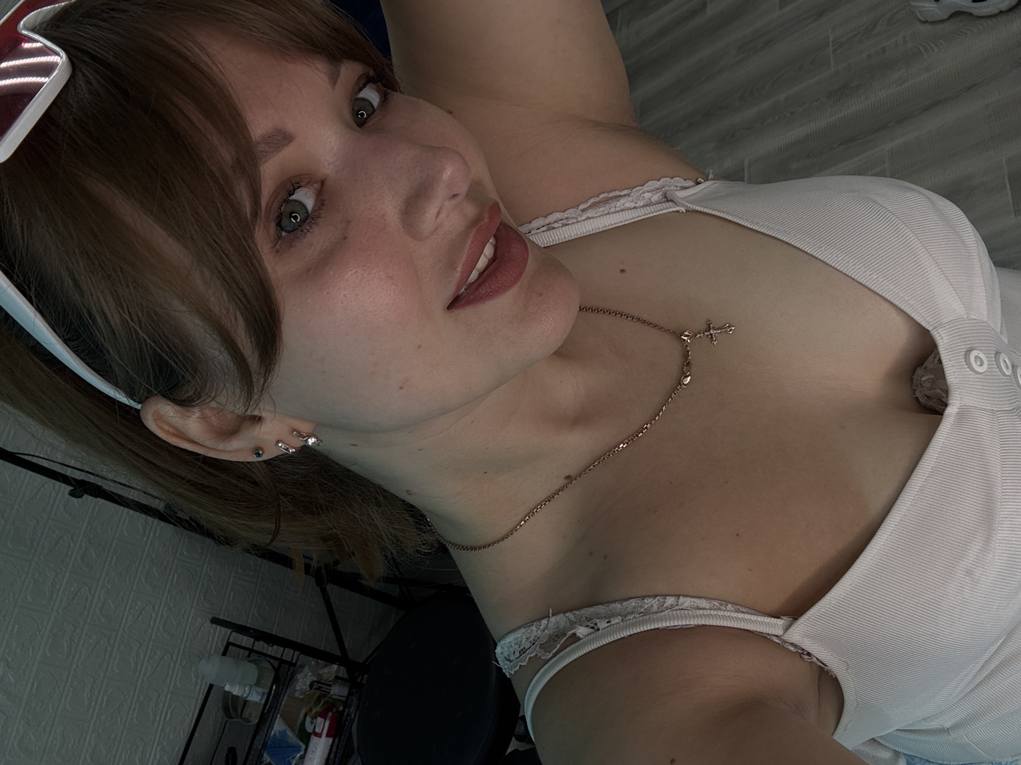 The image is a close-up, selfie-style portrait of a young woman. She has light skin with freckles, green eyes, and light brown hair pulled back. She is wearing a white tank top with lace trim and a rose gold necklace with a cross pendant. She is also wearing sunglasses pushed up onto her head. Her makeup includes mascara and pink lipstick. The background appears to be an indoor setting, with a brick-patterned wall on the left and a wooden floor on the right. There are some objects on a shelf in the background, including bottles and what appears to be a lamp. There are no visible clues that indicate the image was taken in the city of Chaikovsky, Russia.