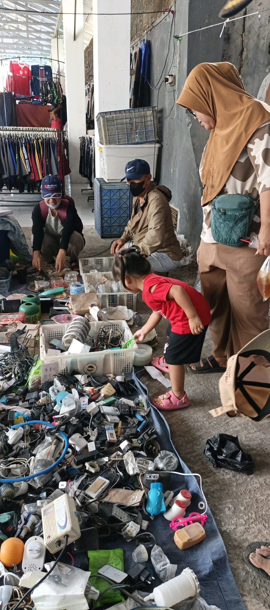 In Jatinom, Indonesia, a group of people are gathered around a collection of discarded items spread on a blue tarp. In the foreground, a young child in a red shirt and black shorts is actively examining the items. To the left, two adults are crouched down, seemingly engaged in sorting or selling the goods. One of them wears a blue cap and a face mask, while the other has a maroon backpack. To the right, an adult in a brown headscarf and a camouflage-patterned shirt looks on, holding a plastic bag. The environment appears to be an outdoor market, with racks of clothing visible in the background. The discarded items are a mix of electronics, cables, adapters, and various household objects.